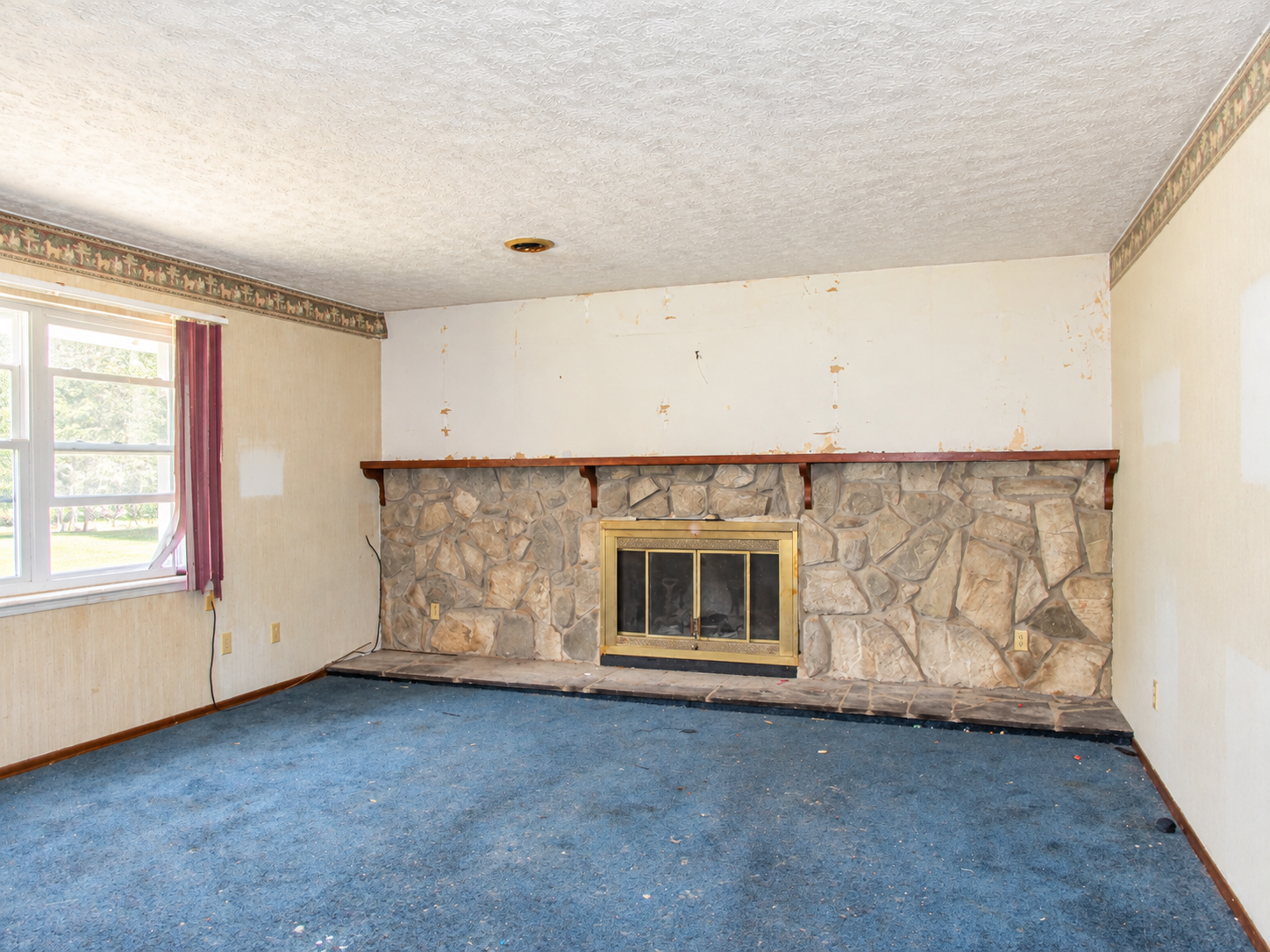 Empty living room with stone fireplace, blue carpet, and a window with red curtains.