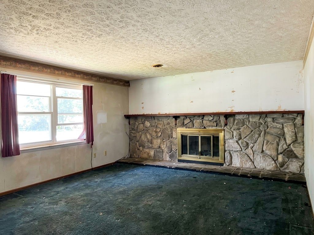 Empty living room with stone fireplace, green carpet, and a window with burgundy curtains