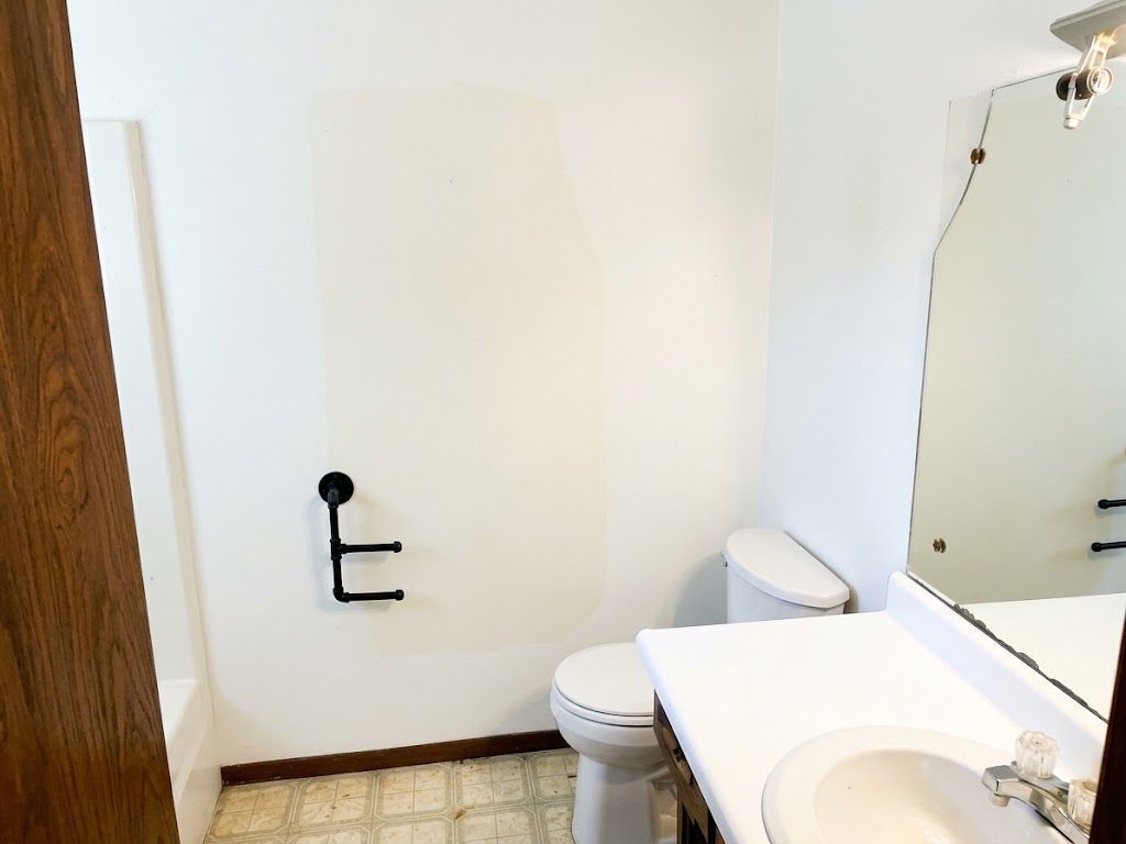 Small bathroom with white walls, toilet, sink, mirror, and grab bar near a wooden door.