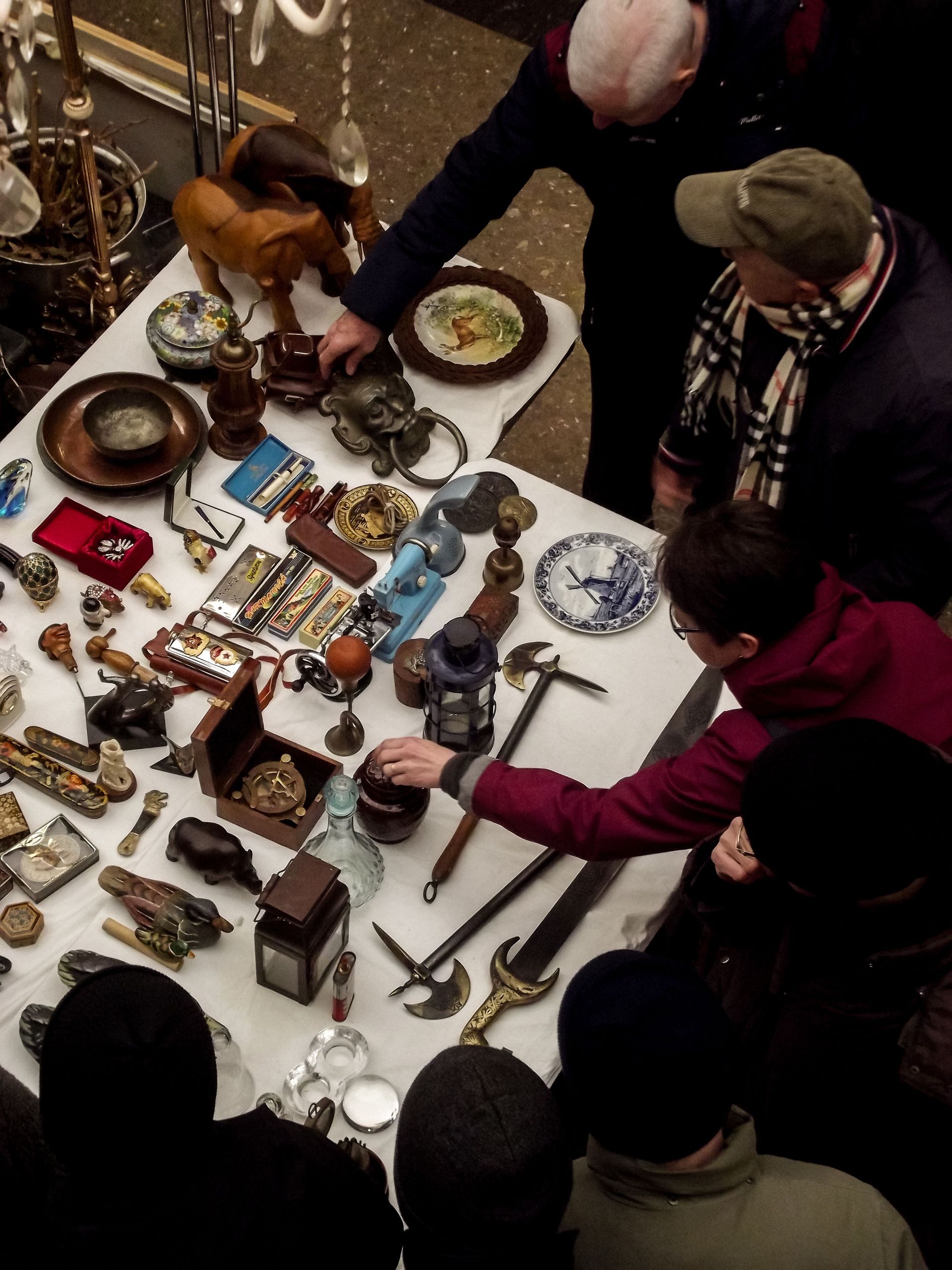 People browsing assorted antiques and jewelry displayed on a table at an indoor market
