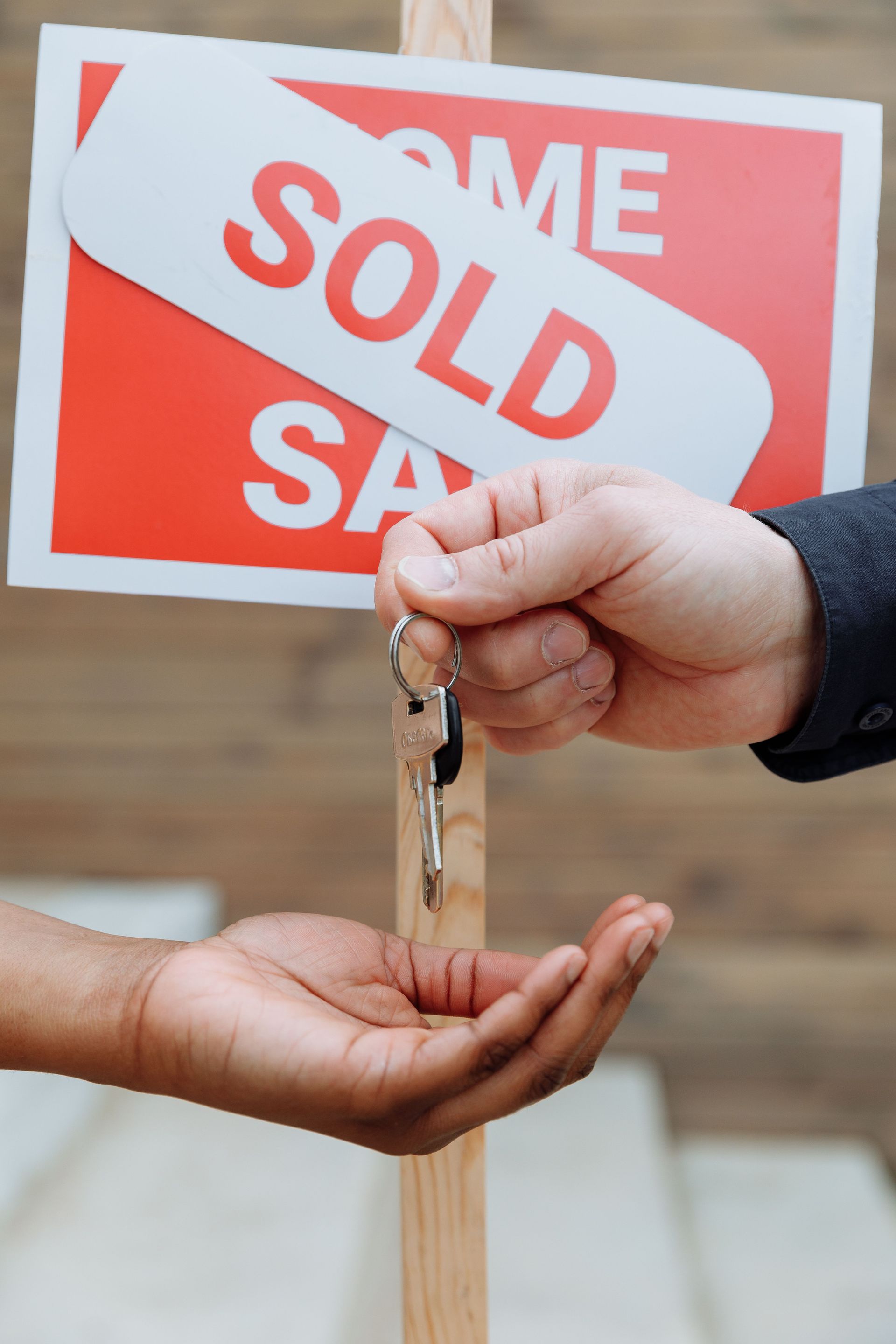 Hands exchanging house keys in front of a red-and-white “SOLD” real estate sign.