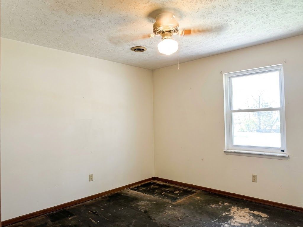 Empty beige room with ceiling fan, window, and worn floor; no furniture