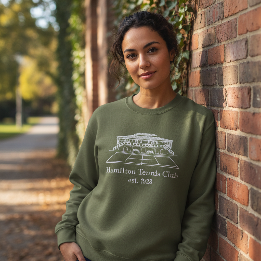 Woman in olive green sweatshirt leans against a brick wall. The sweatshirt has a white logo: Hamilton Tennis Club est. 1921.
