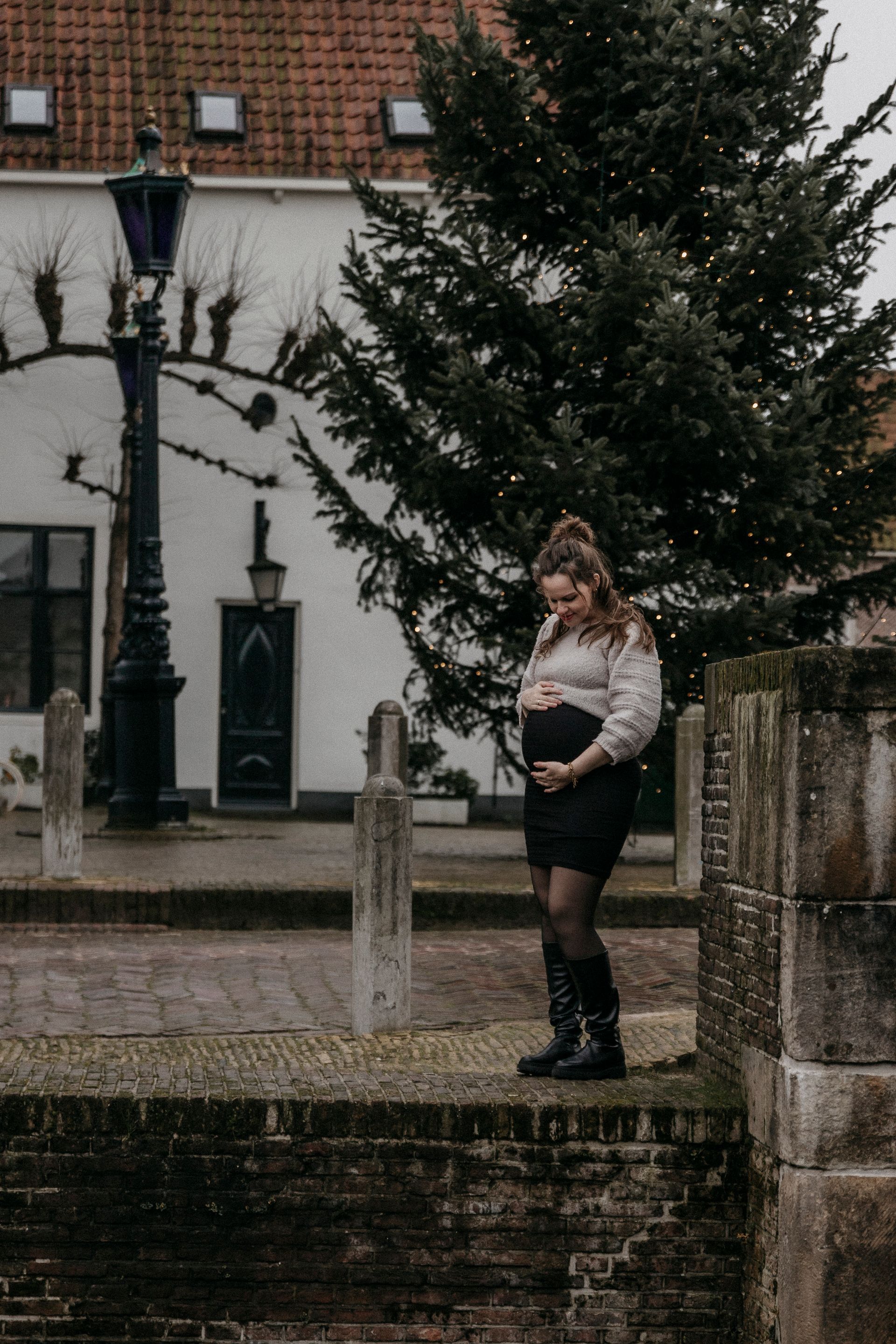 Zwangere vrouw in een zwarte jurk en laarzen buiten, die haar buik aanraakt.