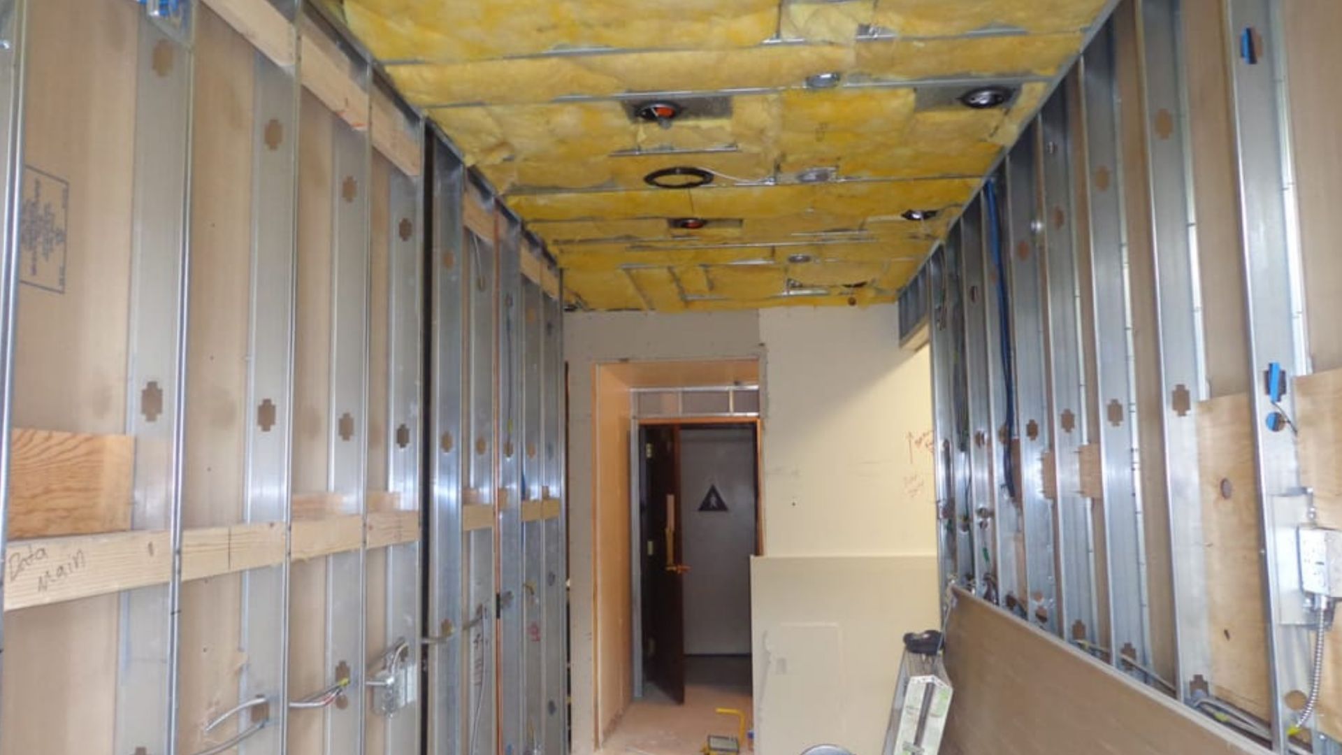 Interior of a narrow room under construction, with exposed wall studs, insulation, and a yellow ceiling.