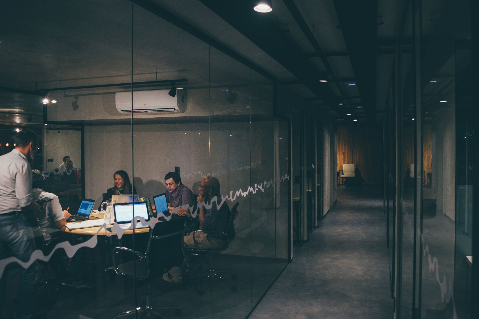 Office meeting in glass-walled room; people working on laptops; dim lighting, hallway visible.