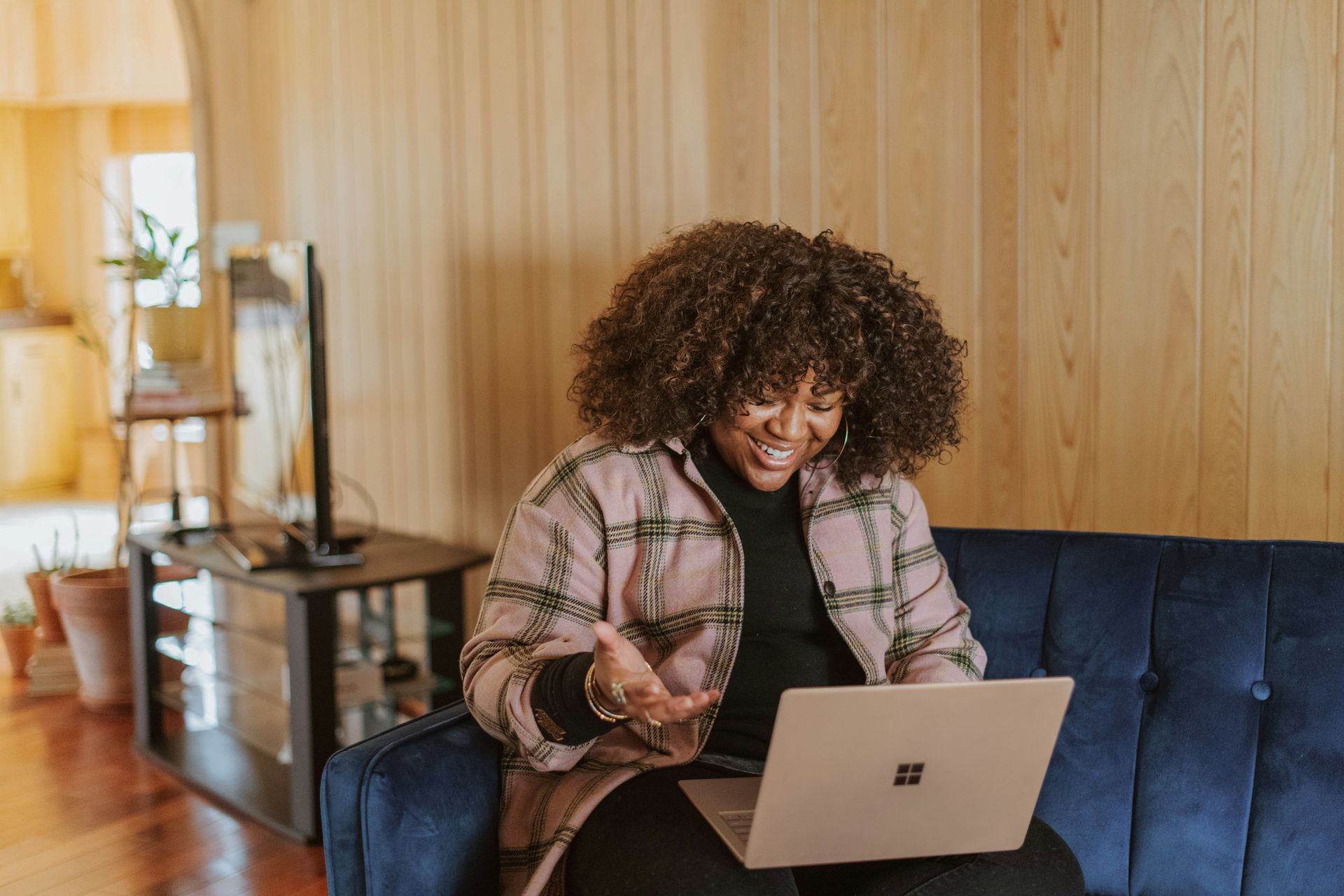 Woman with curly hair smiles at laptop on blue couch. Indoors with wood paneling.