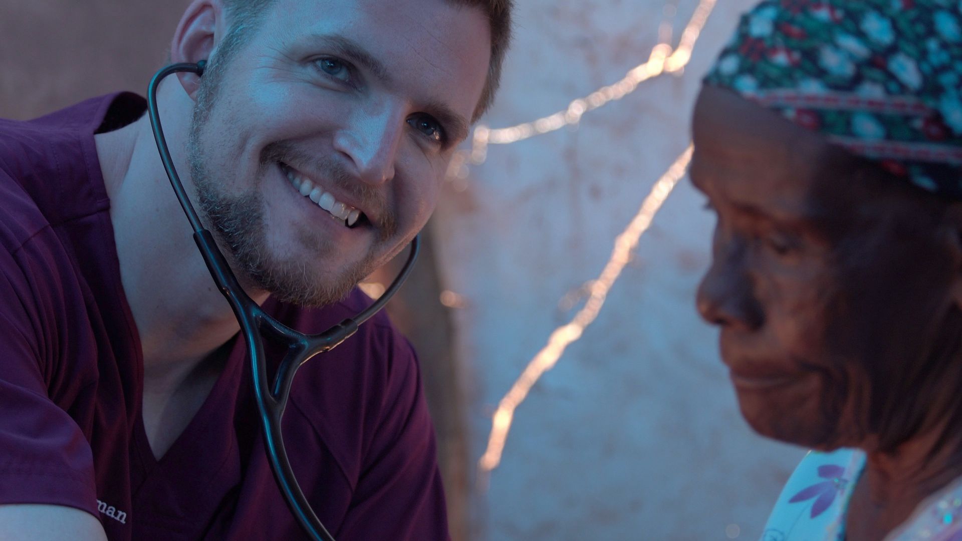 A smiling doctor in maroon scrubs listens to a patient's chest with a stethoscope.