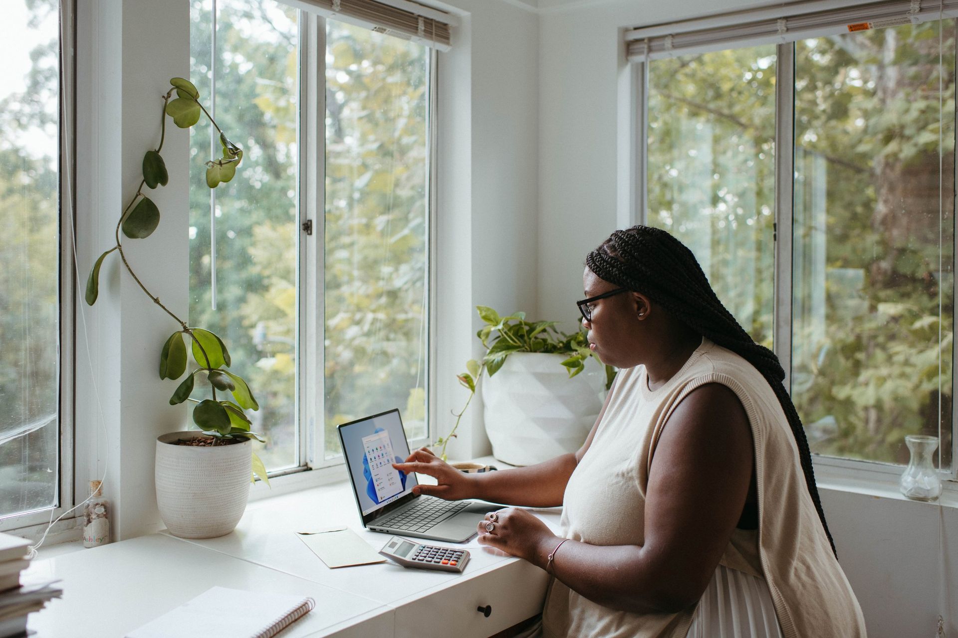 Woman working on laptop near a window with plants; analyzing data.
