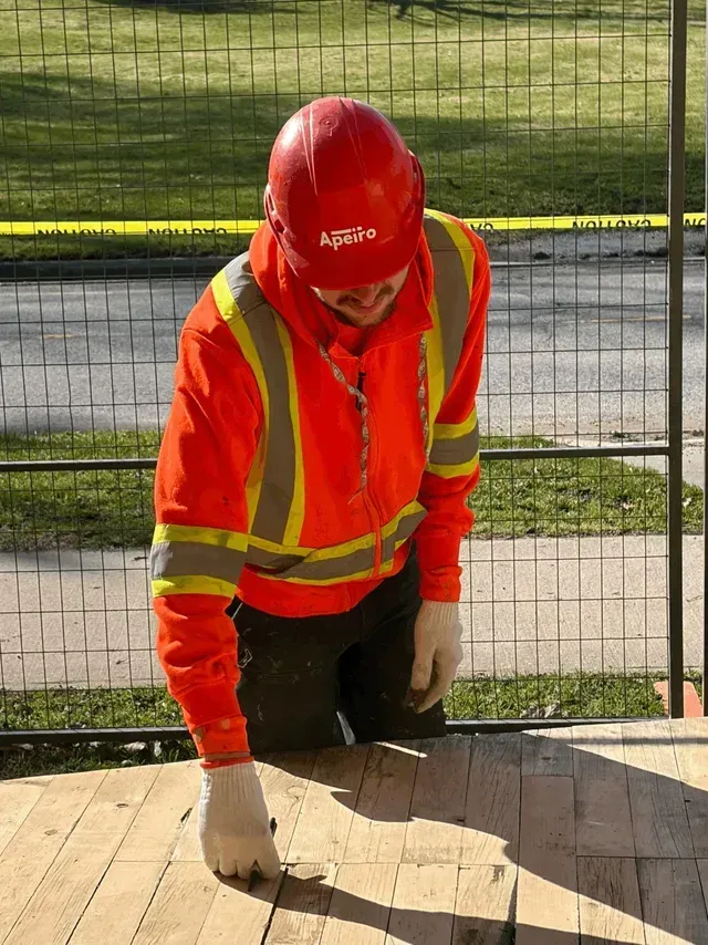 Worker in orange safety gear and red hard hat kneeling beside a wooden deck outdoors.