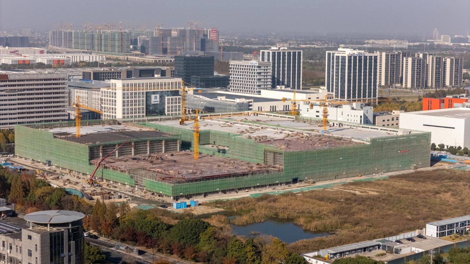 Aerial view of a large construction site with a partially built building, cranes, and surrounding city buildings.