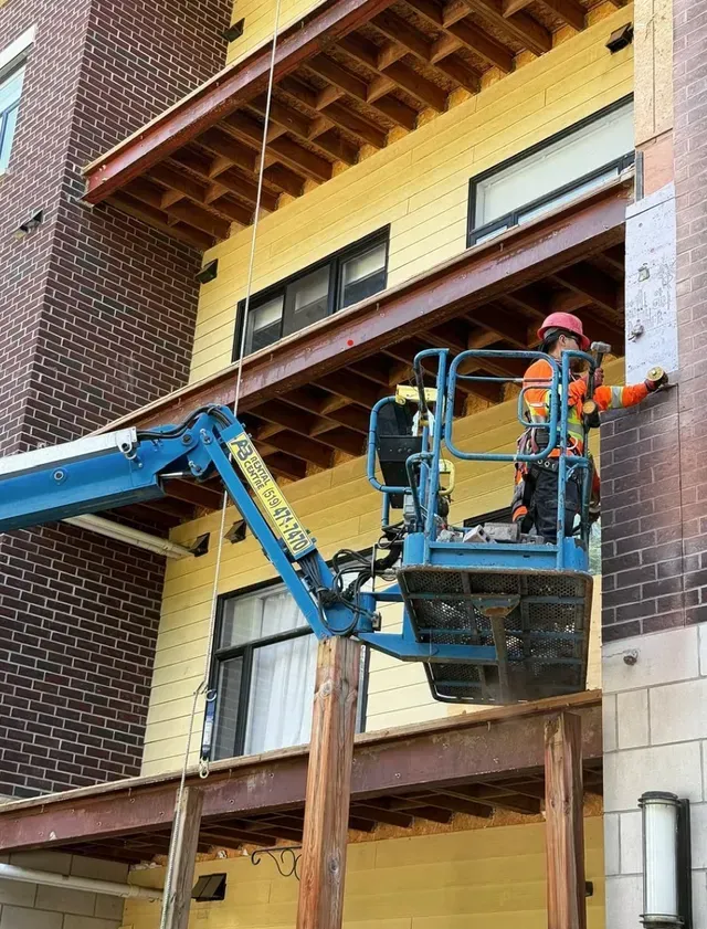 A worker in high-visibility gear stands in a blue lift bucket, reaching to install equipment on a building exterior.