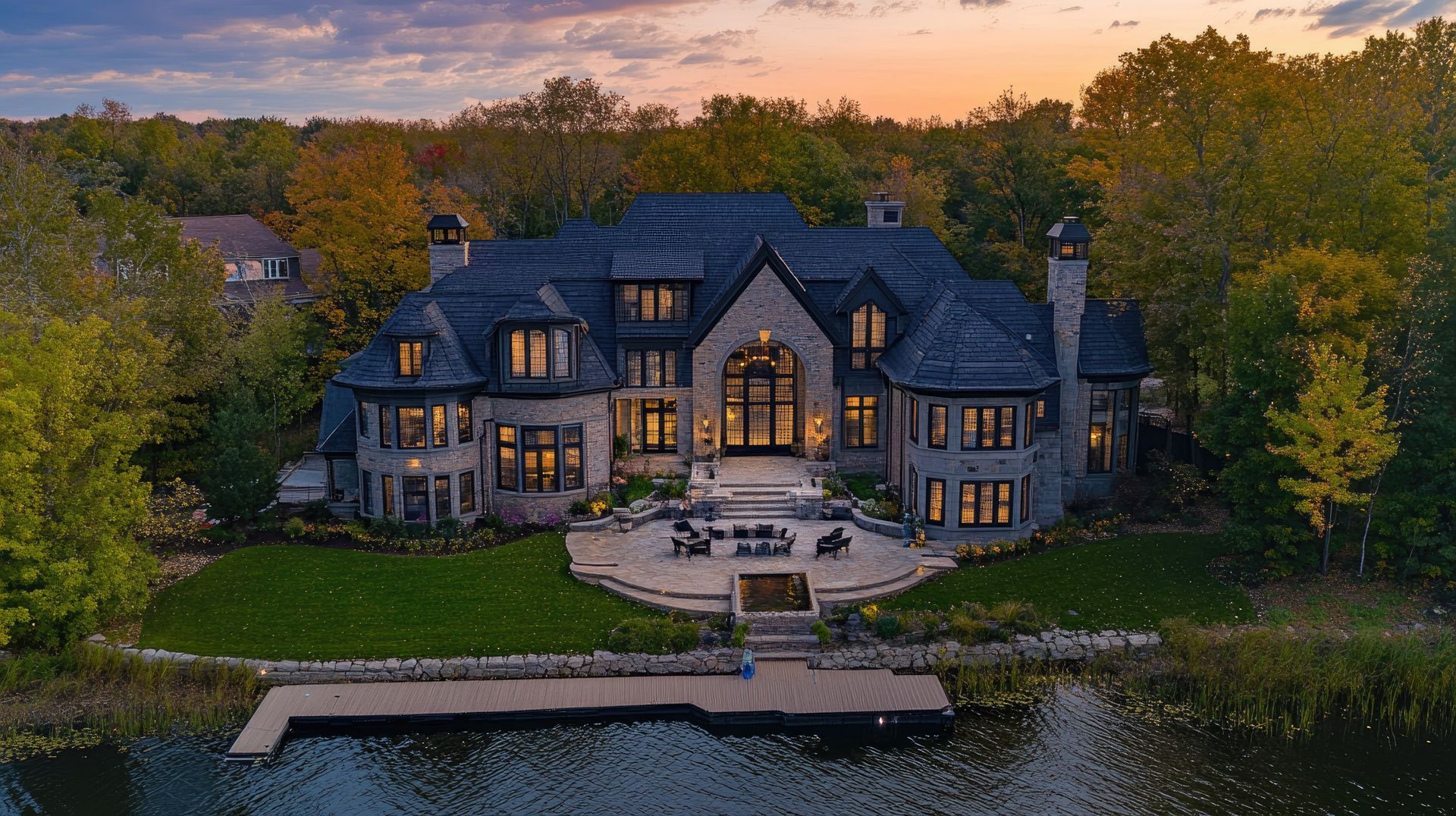 Large lakeside stone house at sunset, with warm lights and autumn trees surrounding it.