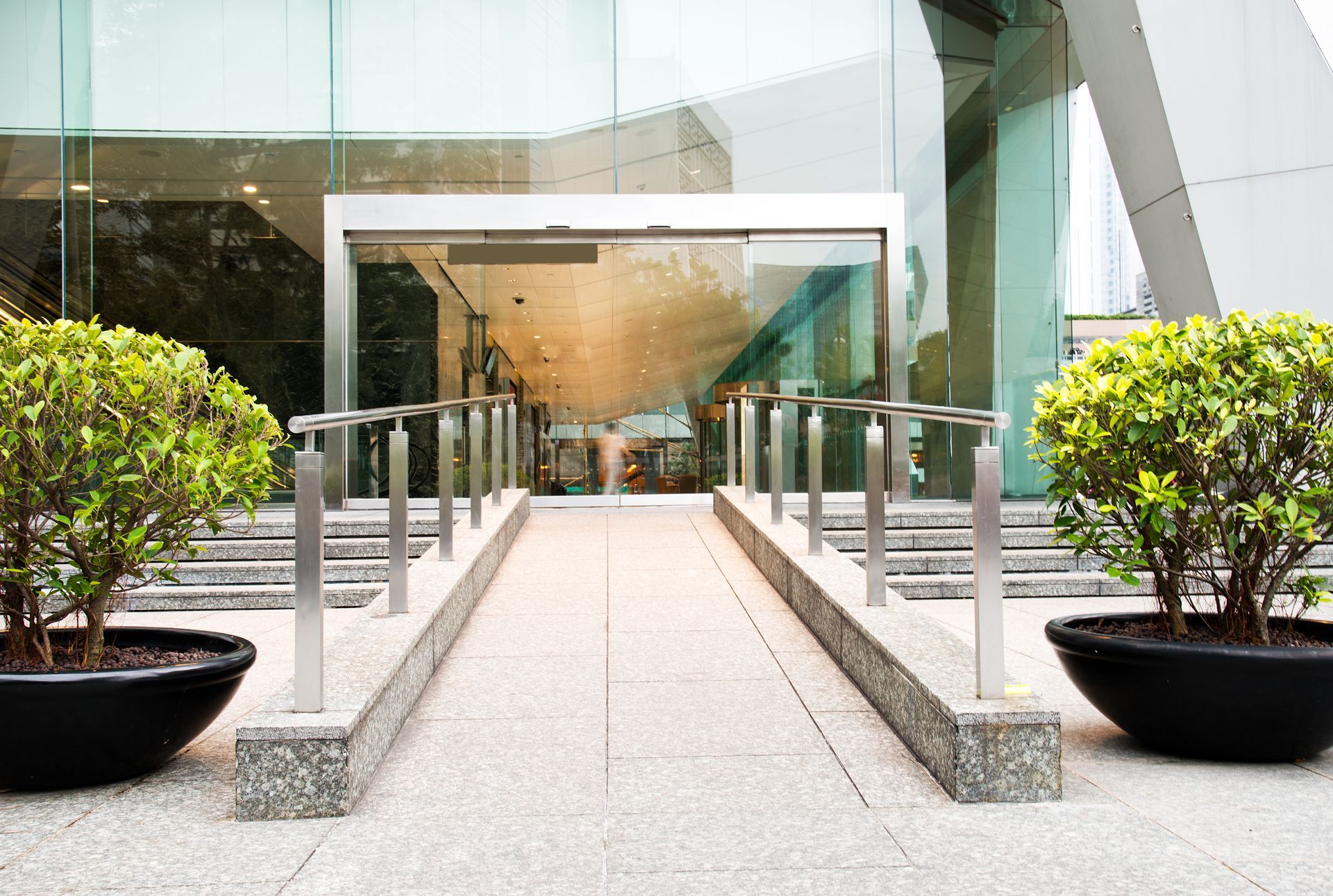 Modern building entrance with glass doors, concrete walkway, and large potted shrubs on both sides