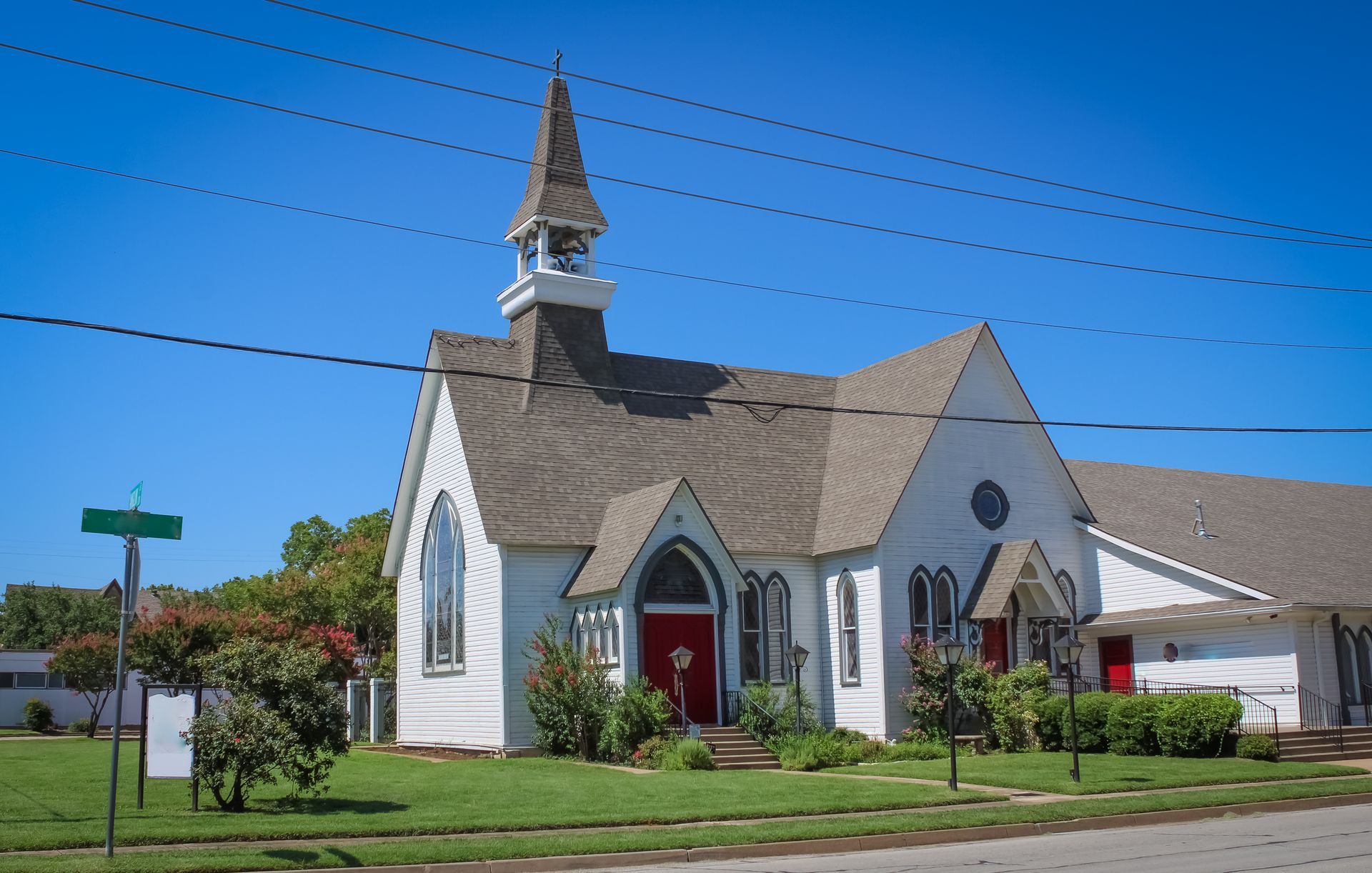 Small white church with a red door and steeple on a sunny street corner