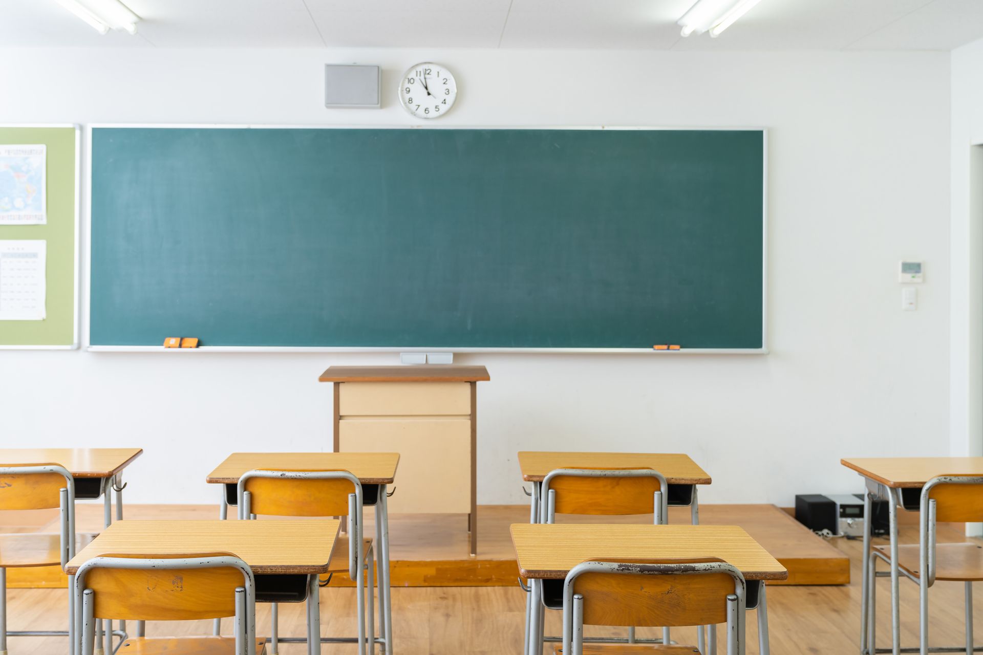Empty classroom with green chalkboard, wooden desks, and a teacher’s podium under wall clock
