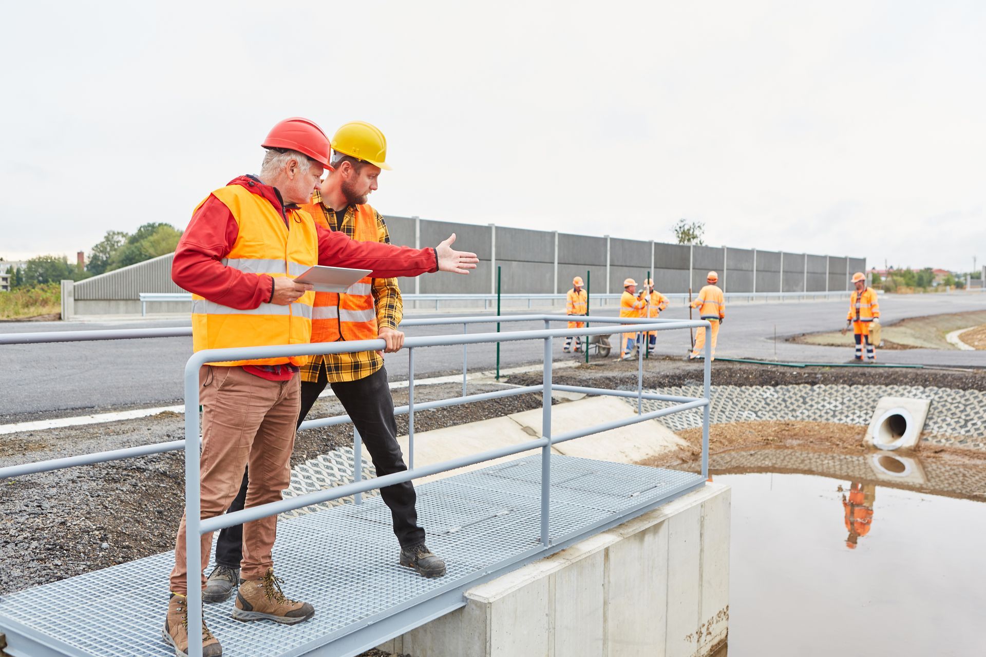 Two workers in hard hats inspect a bridge walkway above water at a construction site