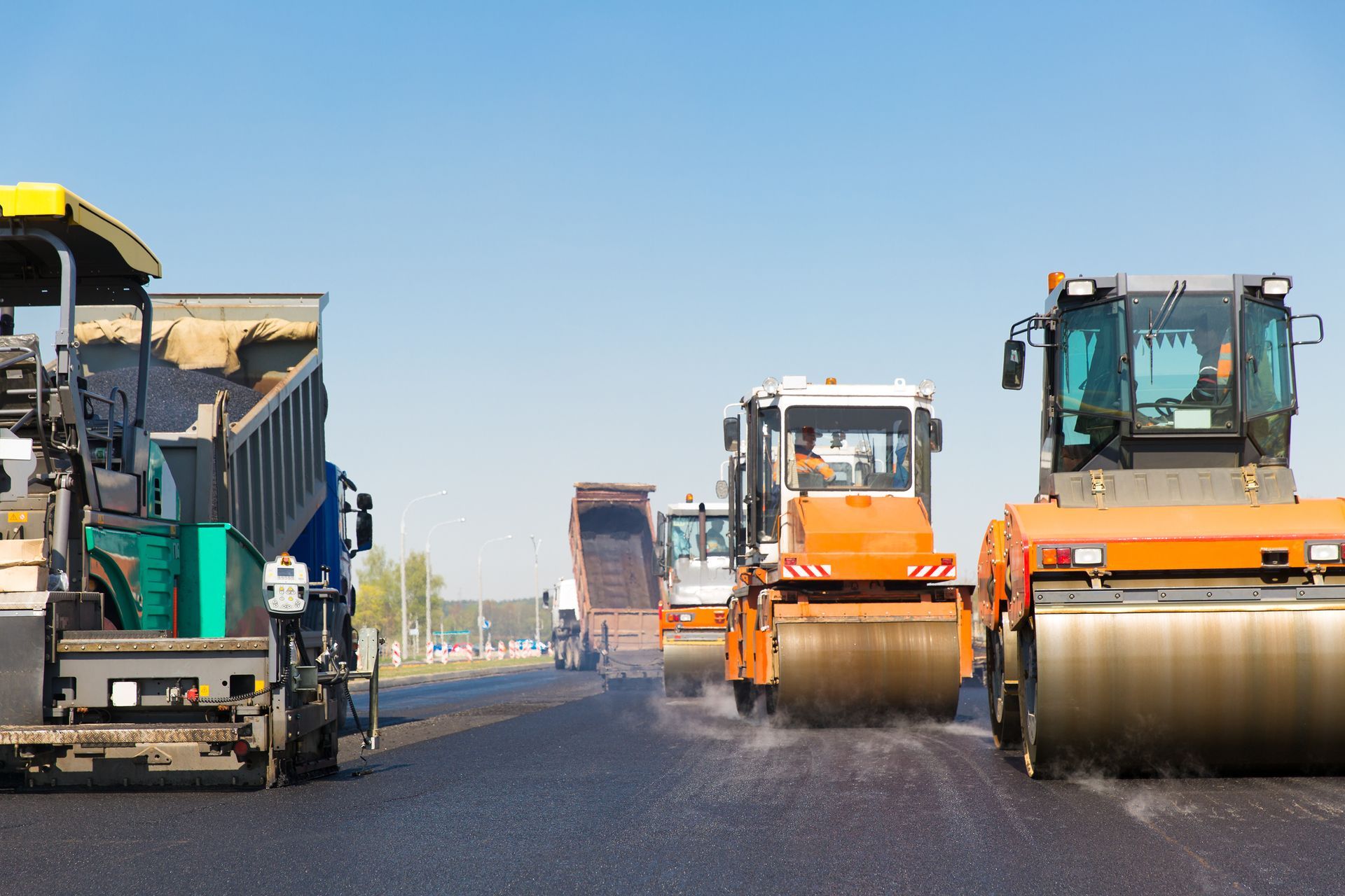 Heavy road rollers paving a roadway, with a yellow asphalt machine on the left and orange rollers on the right