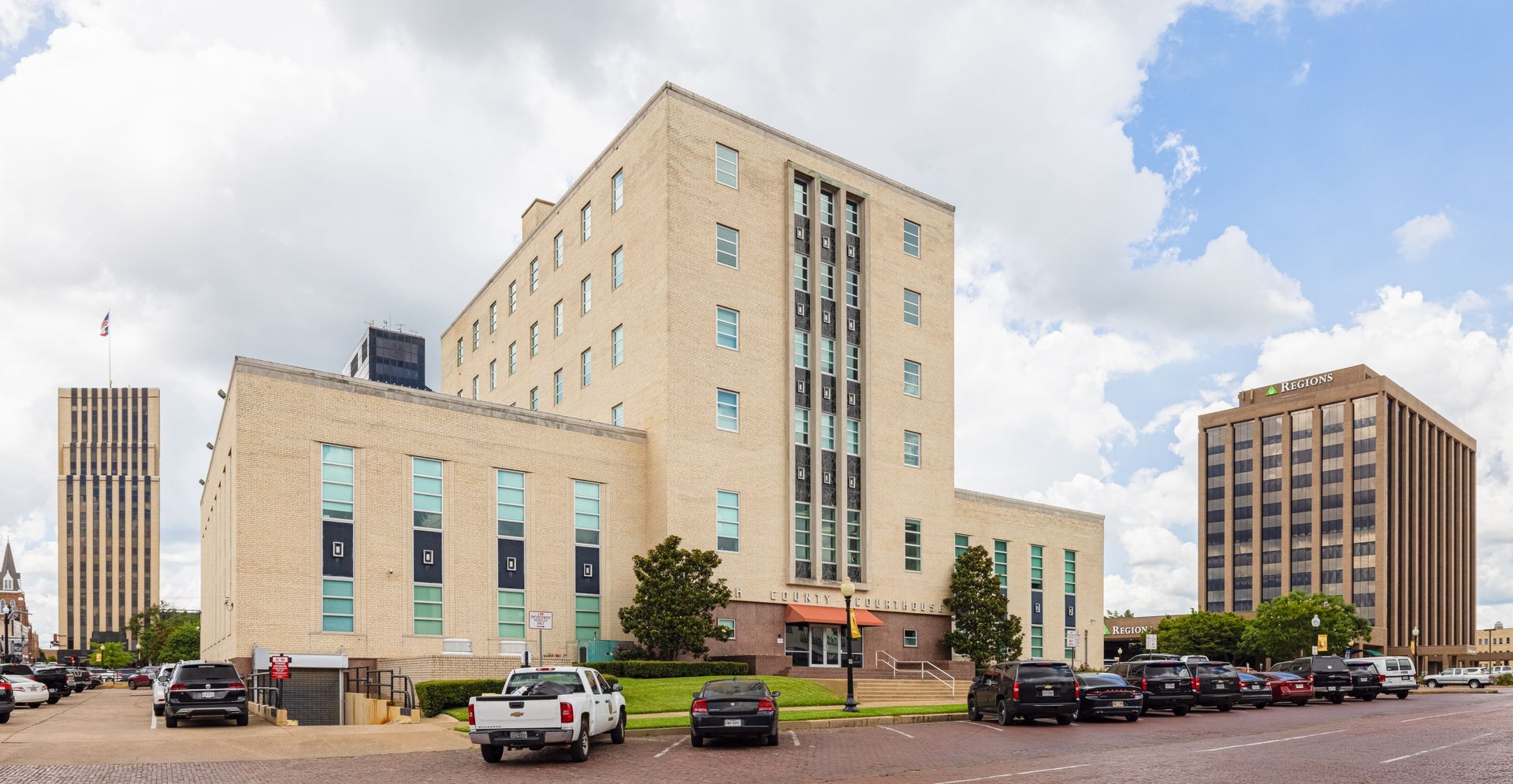 Large beige office building with a parking lot and several cars under a partly cloudy sky