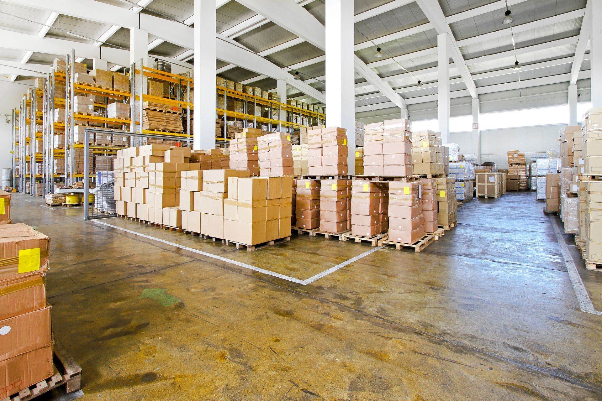 Warehouse with stacked cardboard boxes on pallets, white beams, and a wet concrete floor