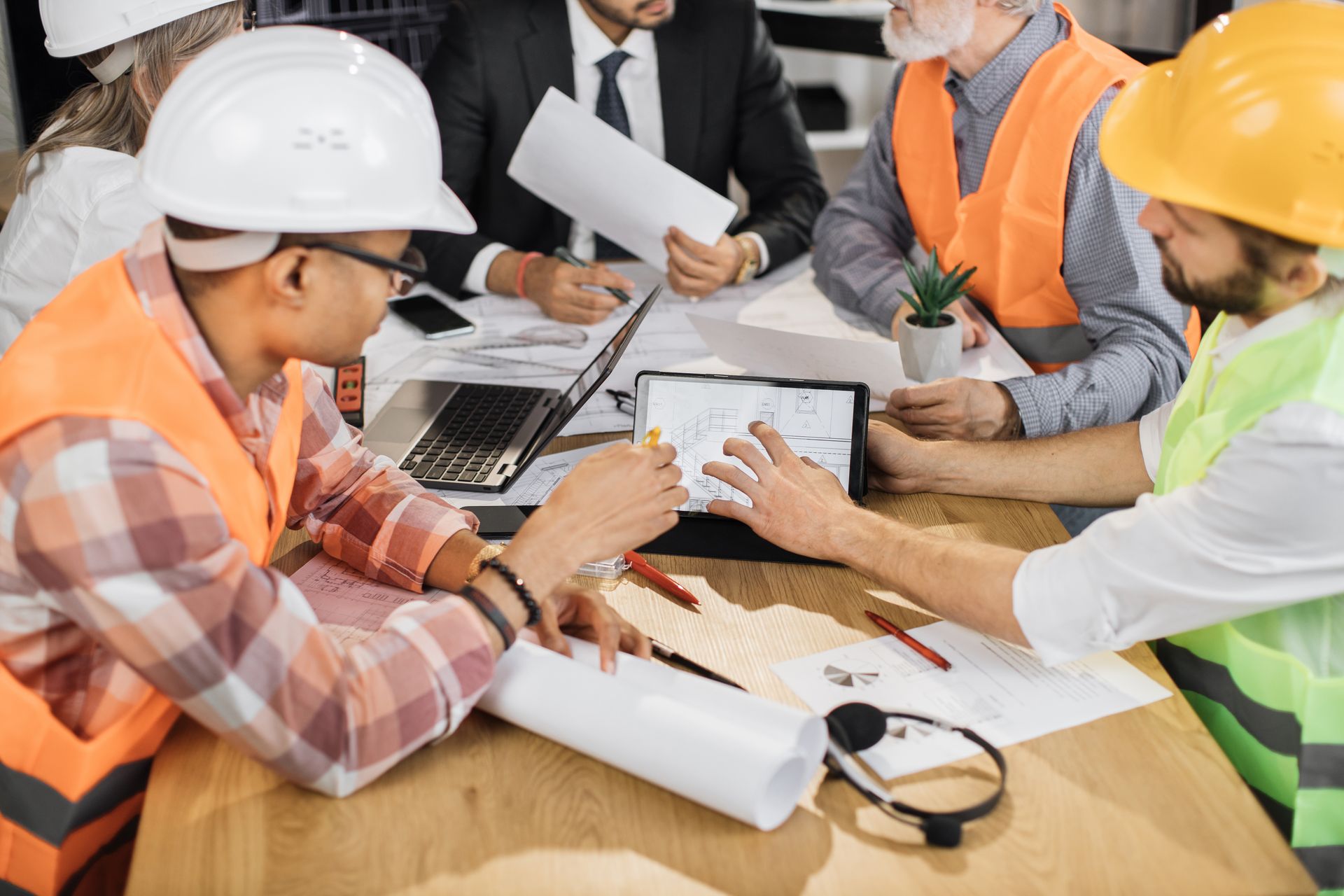 Engineers in hard hats reviewing blueprints and a tablet at a construction meeting