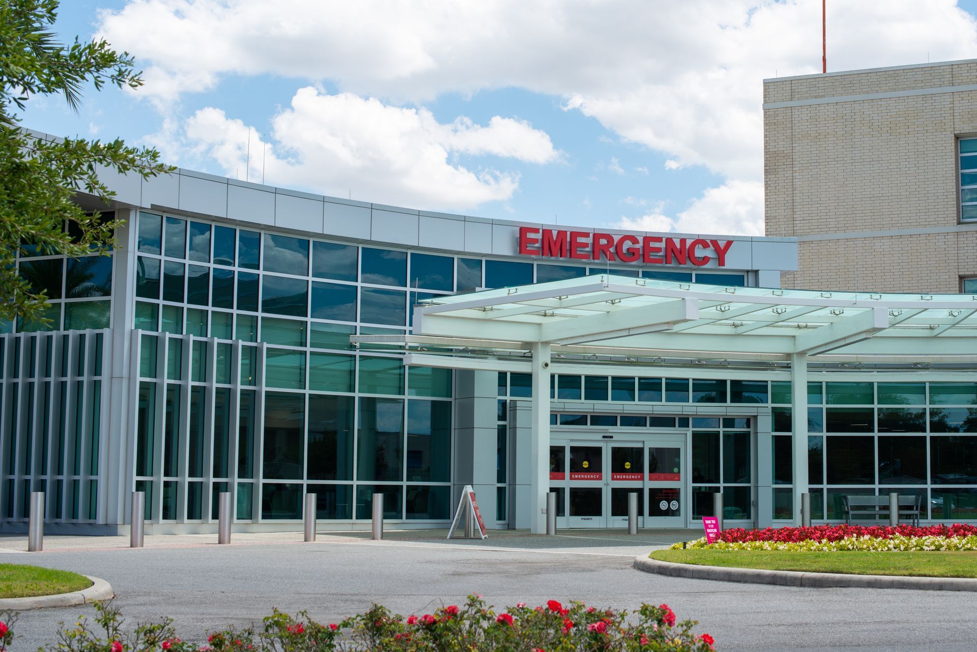 Hospital emergency entrance with glass facade and red “EMERGENCY” sign above a covered walkway.
