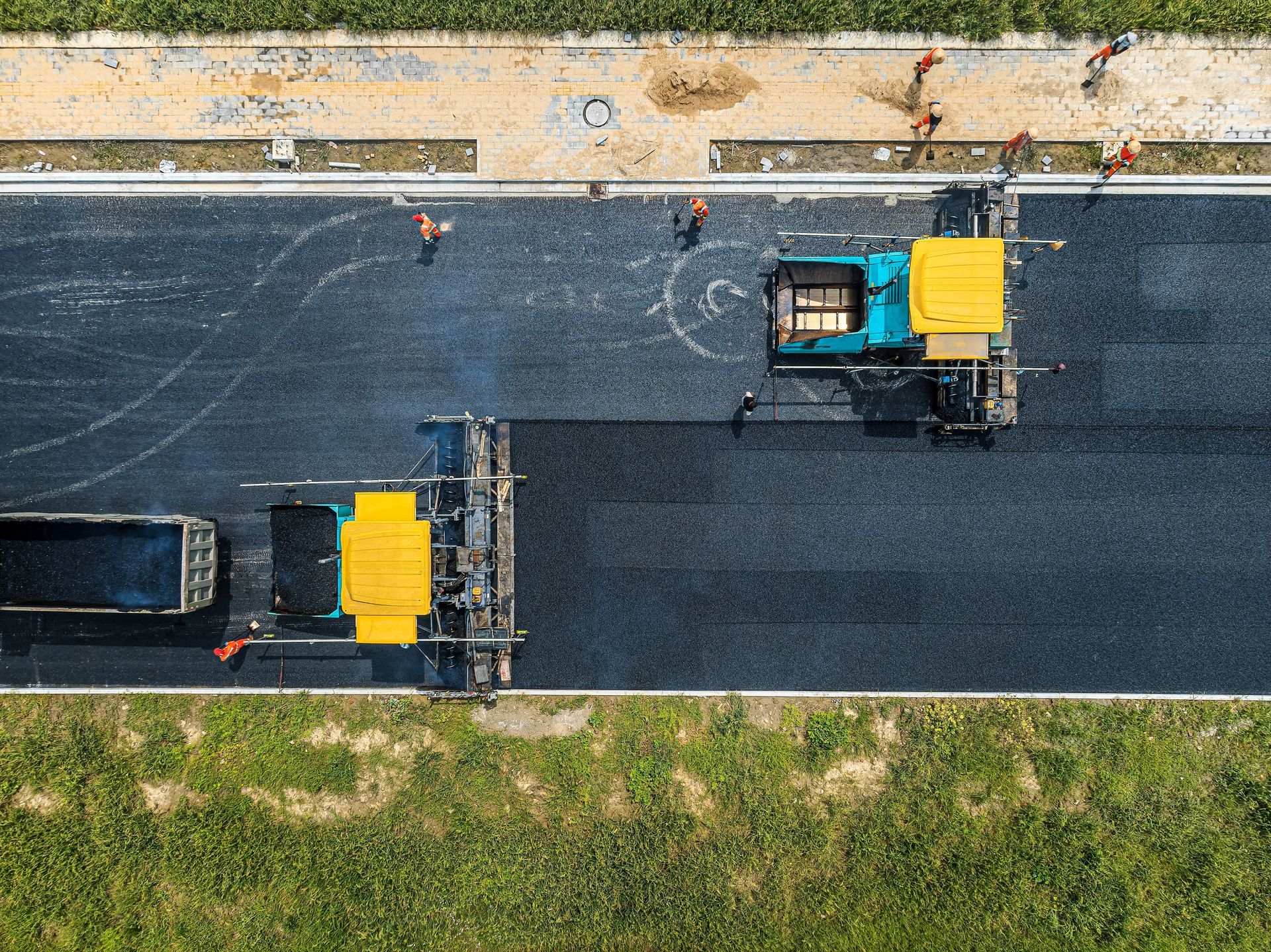 Aerial view of two yellow roadwork machines paving a dark road beside grass and a sidewalk.