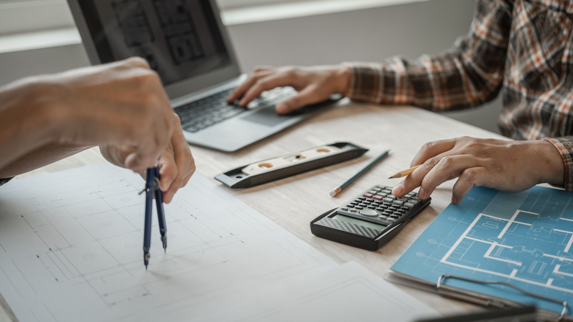Hands working over a desk with laptop, calculator, ruler, and blueprints during a planning meeting
