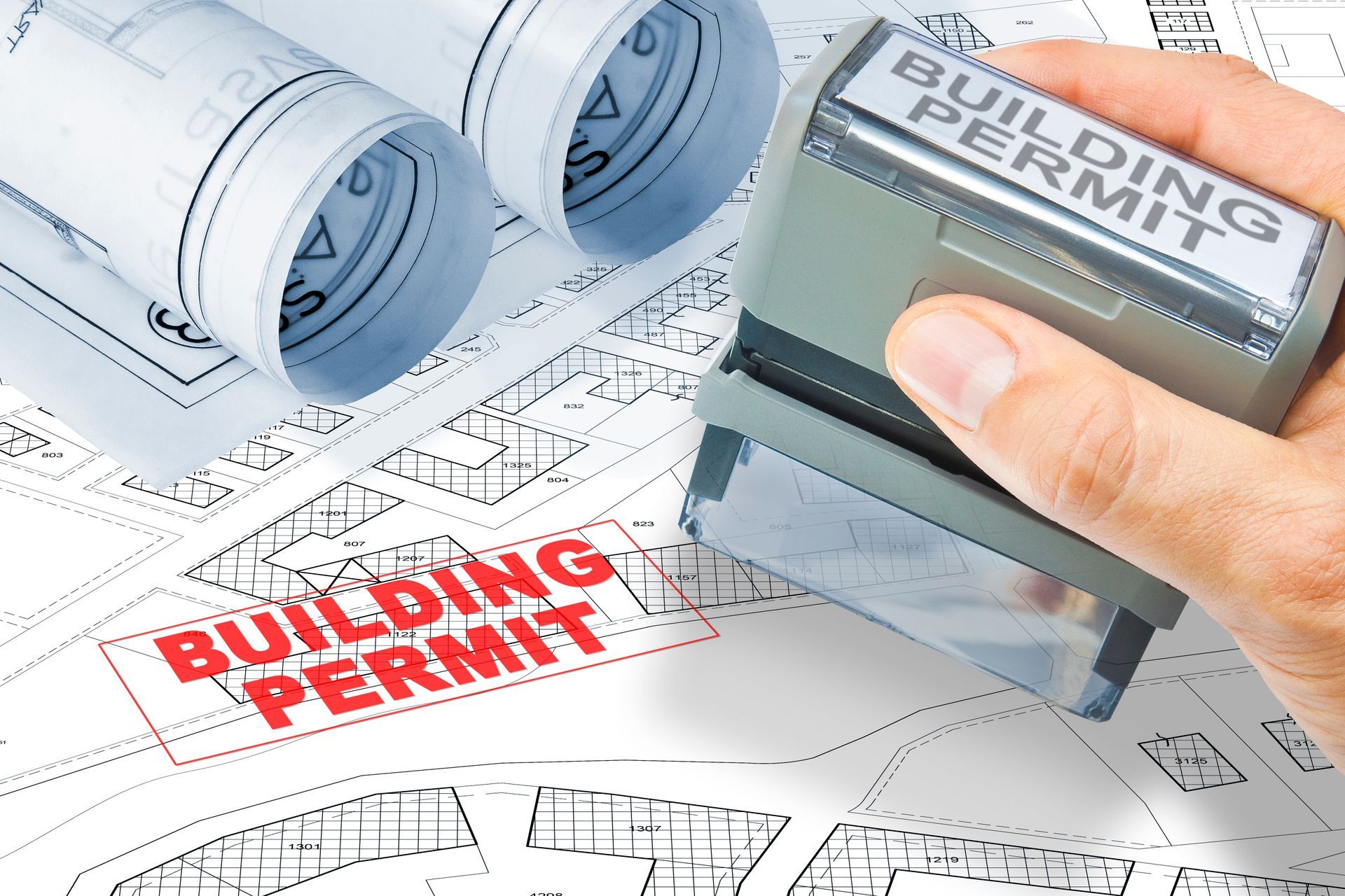 Hand stamping “Building Permit” on architectural blueprints with rolled plans nearby
