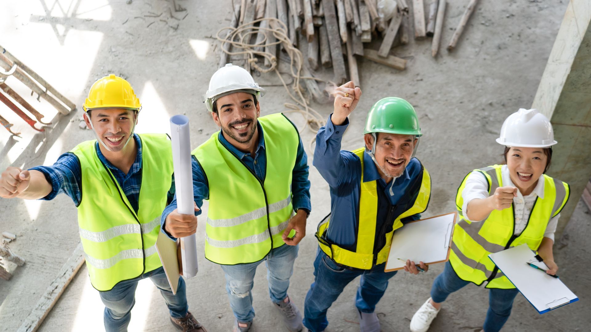 Four construction workers in hard hats and safety vests posing on a building site, smiling and waving up at the camera