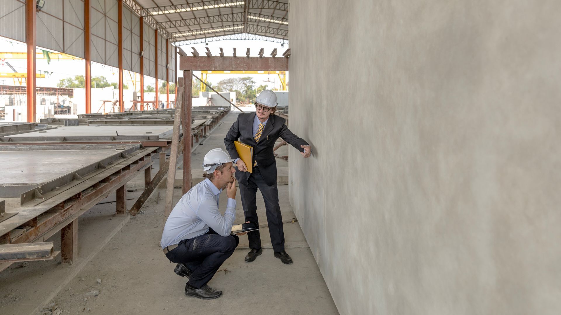 Two construction workers in hard hats inspect a large concrete wall inside a building under construction.