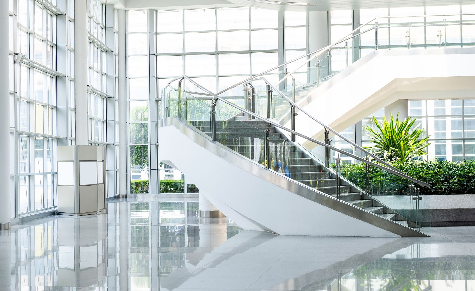 Bright modern atrium with glass walls, white staircase, polished floors, and indoor plants