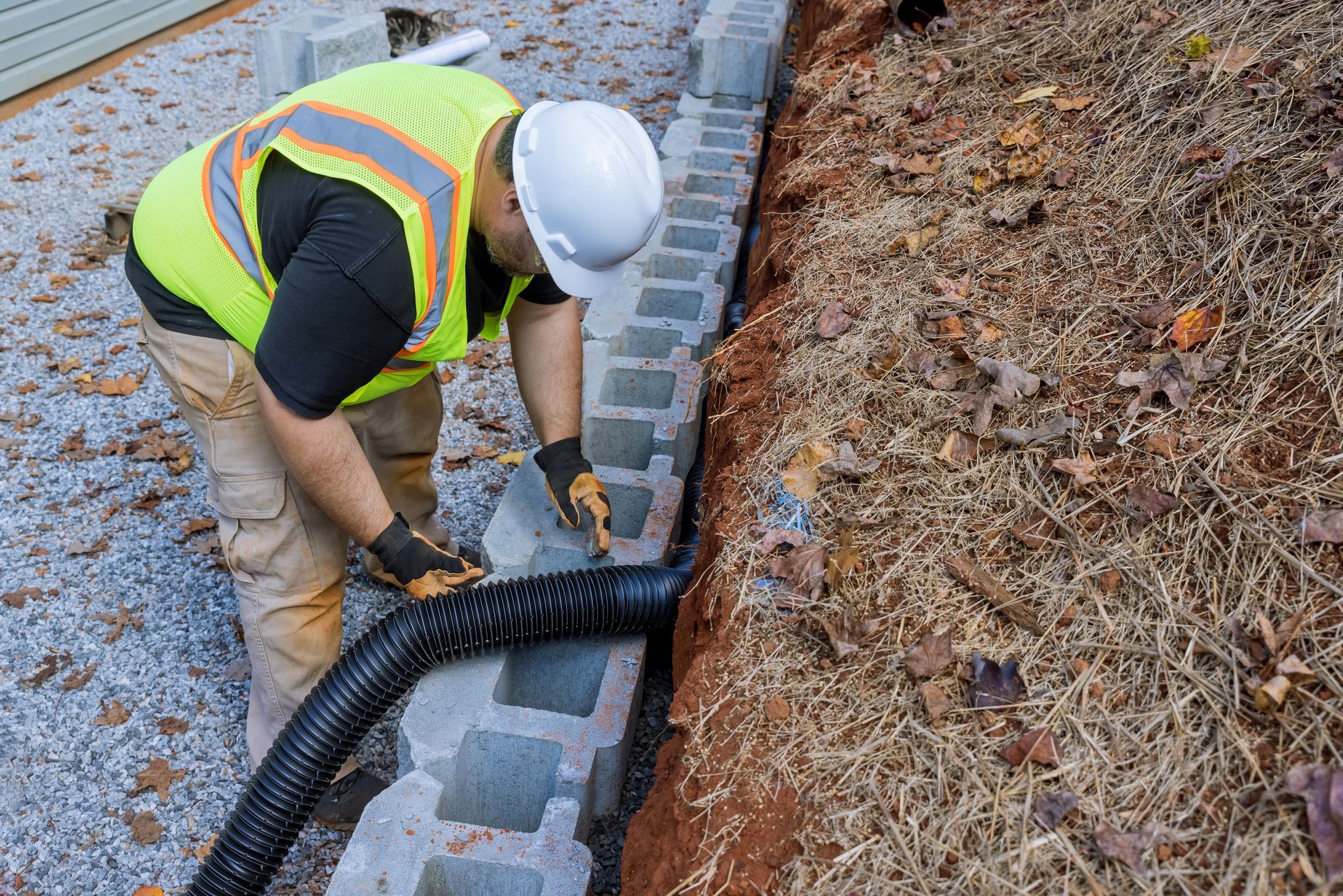 Worker installing black drain pipe along roadside curb beside a gravel and mulch embankment