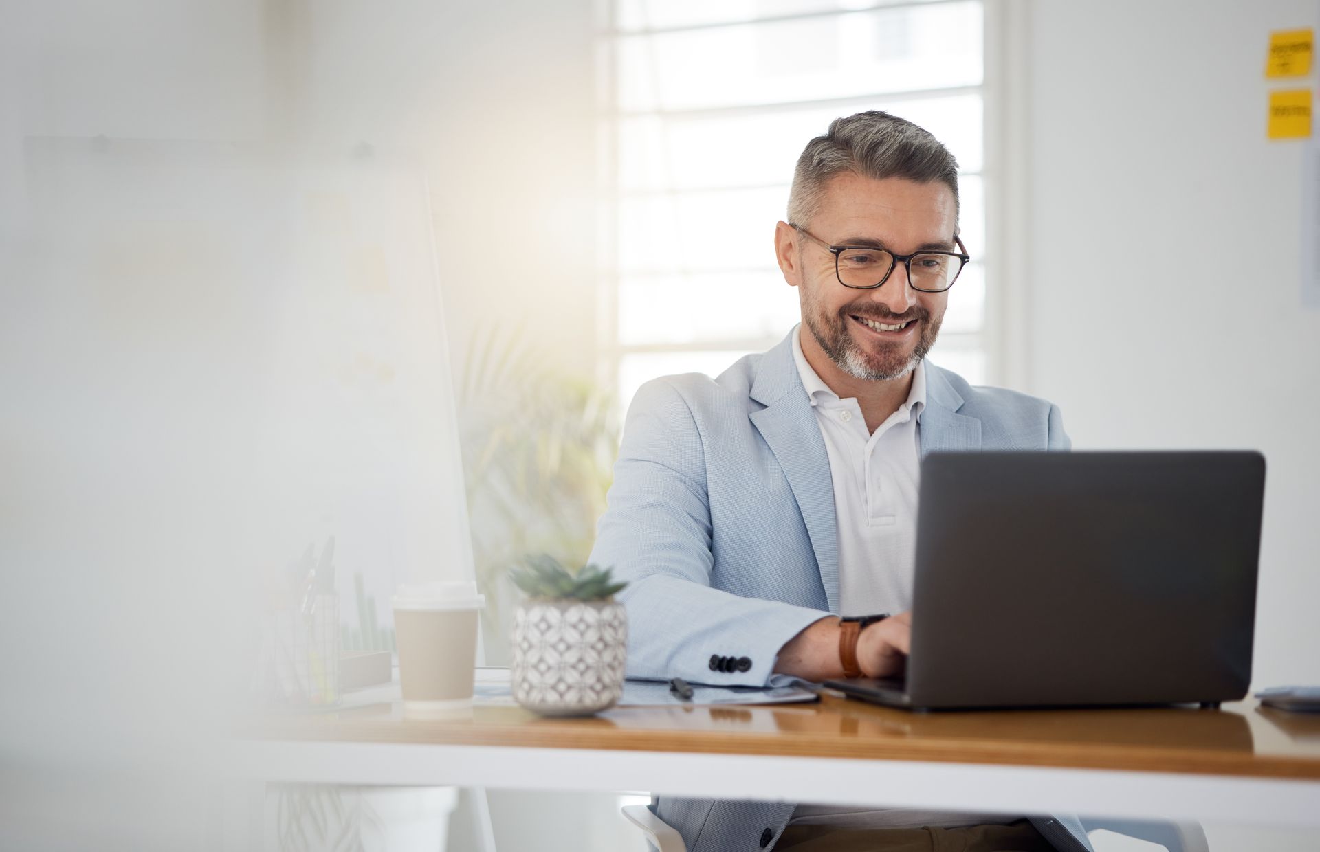 Smiling man in a light blue blazer works on a laptop at a bright desk by a window