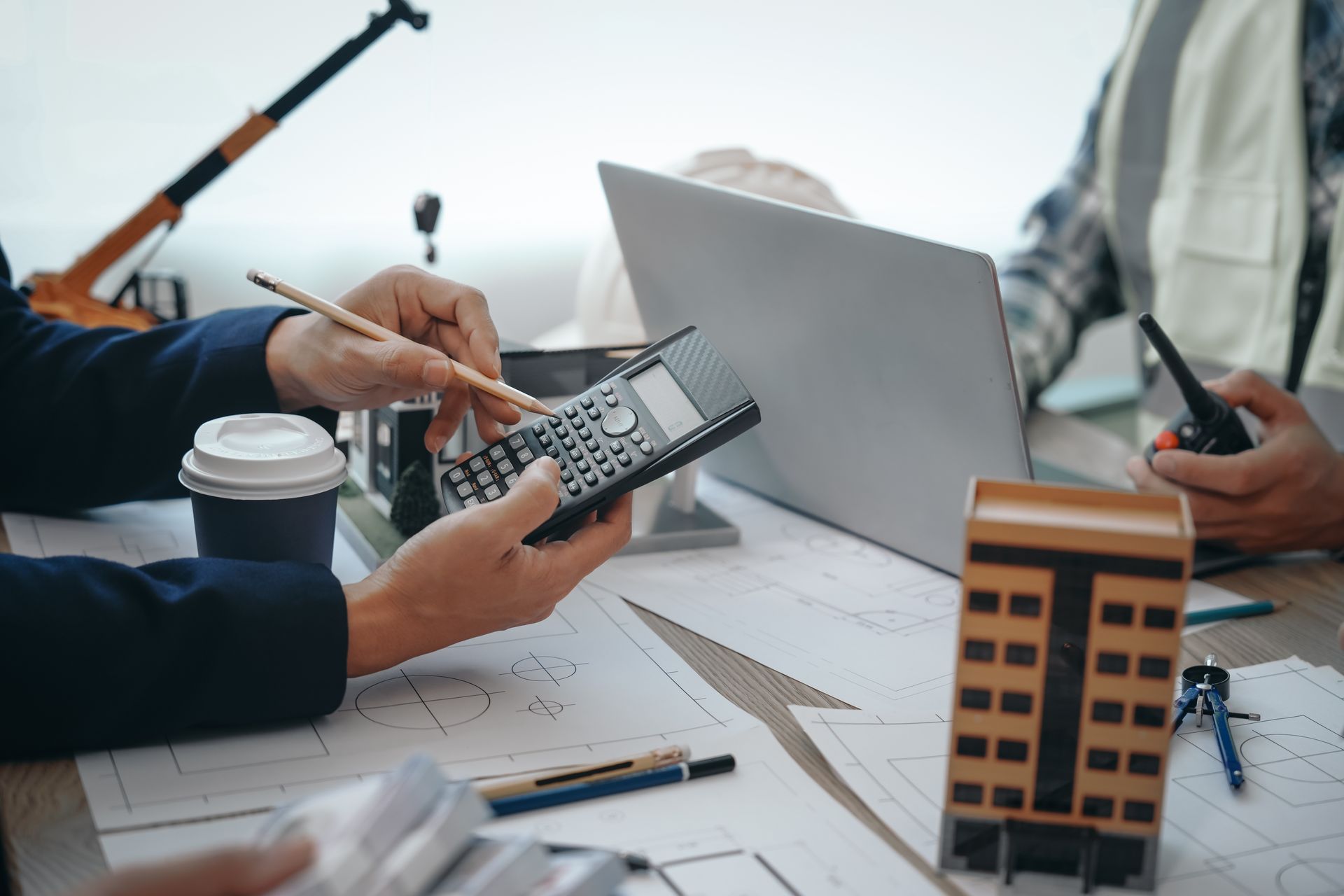 Person using a calculator at a desk with laptop, coffee, and architectural model plans