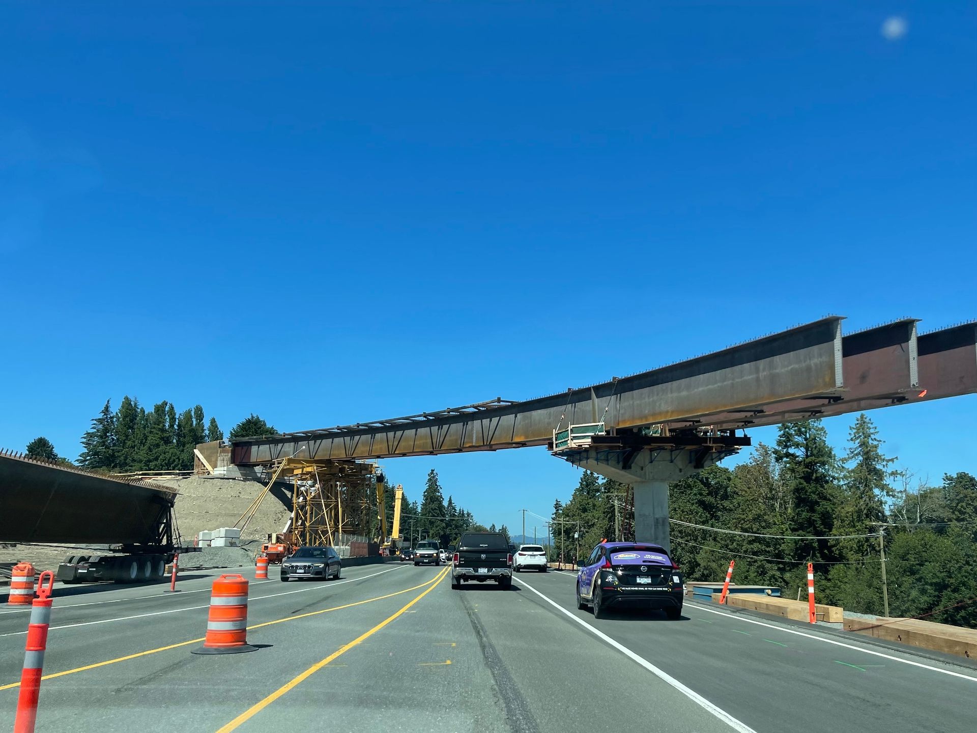 Highway under construction with orange cones and a large overpass beam being installed above traffic
