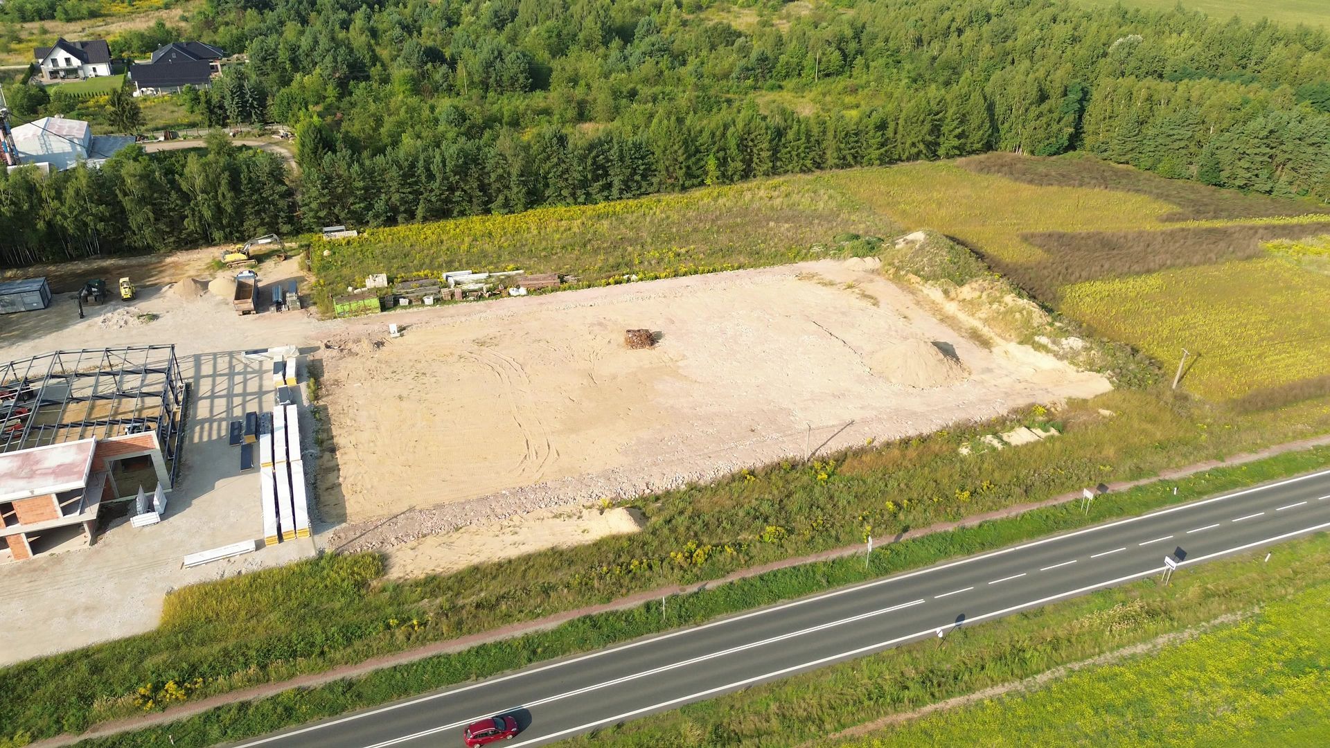 Aerial view of a large sandy construction site beside a road and green fields, with forest in the background