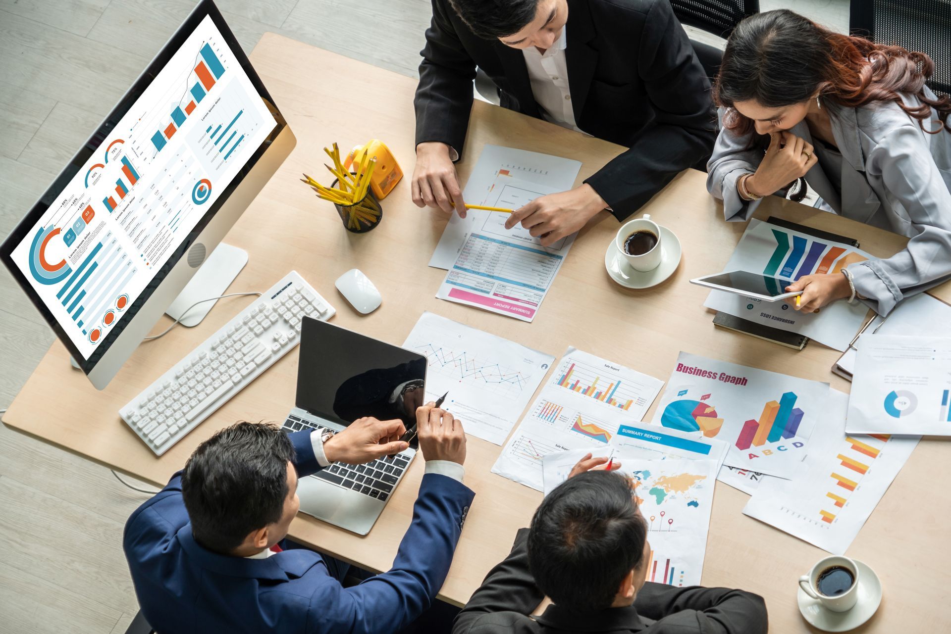 Team meeting around a desk with charts, laptops, and a large monitor displaying analytics graphs.