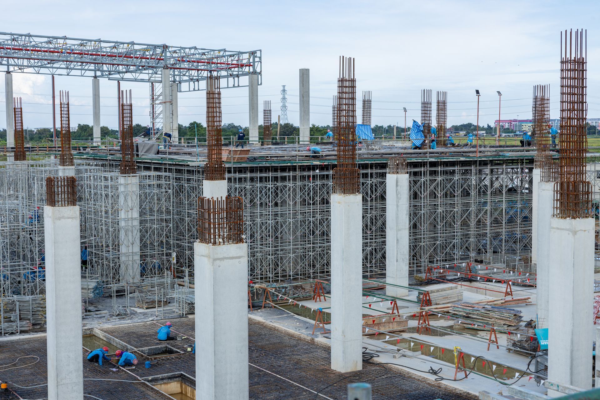 Construction site with concrete columns, steel framework, and workers in blue helmets under an overcast sky