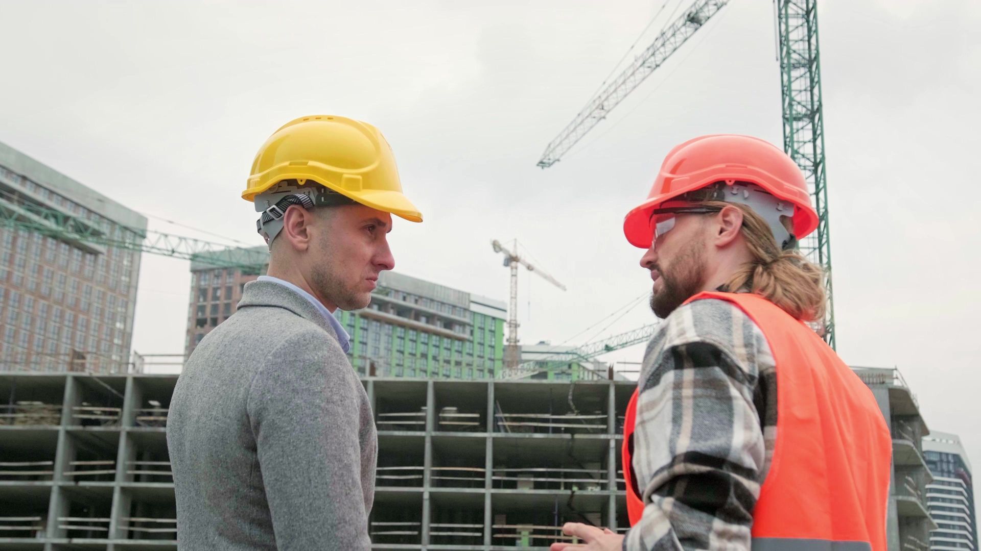 Two construction workers in hard hats talking at a building site, with cranes and a partly built structure behind them.