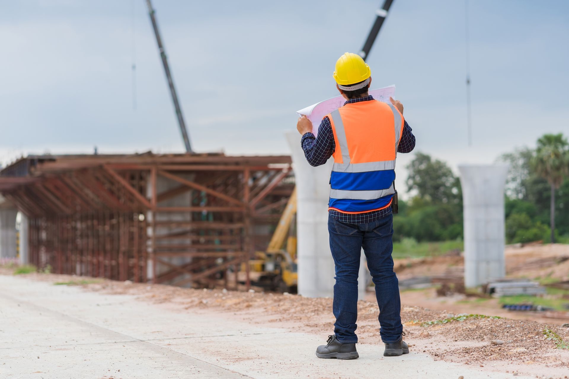 Construction worker in orange safety vest and hard hat standing near a bridge under construction.