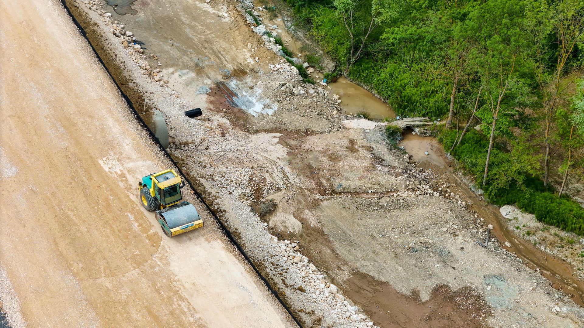 A bulldozer works on a muddy road beside a washed-out embankment and trees.