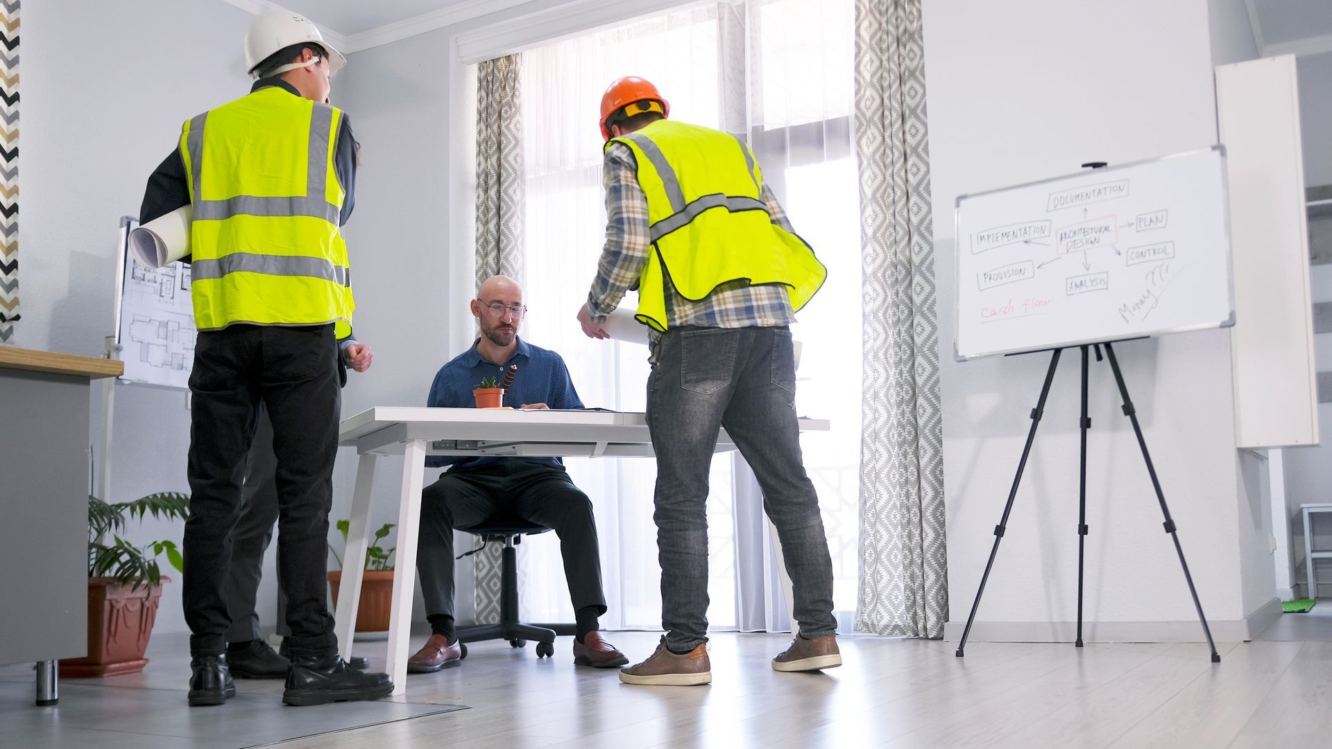 Three workers in high-visibility vests discuss plans in a bright office with a whiteboard.