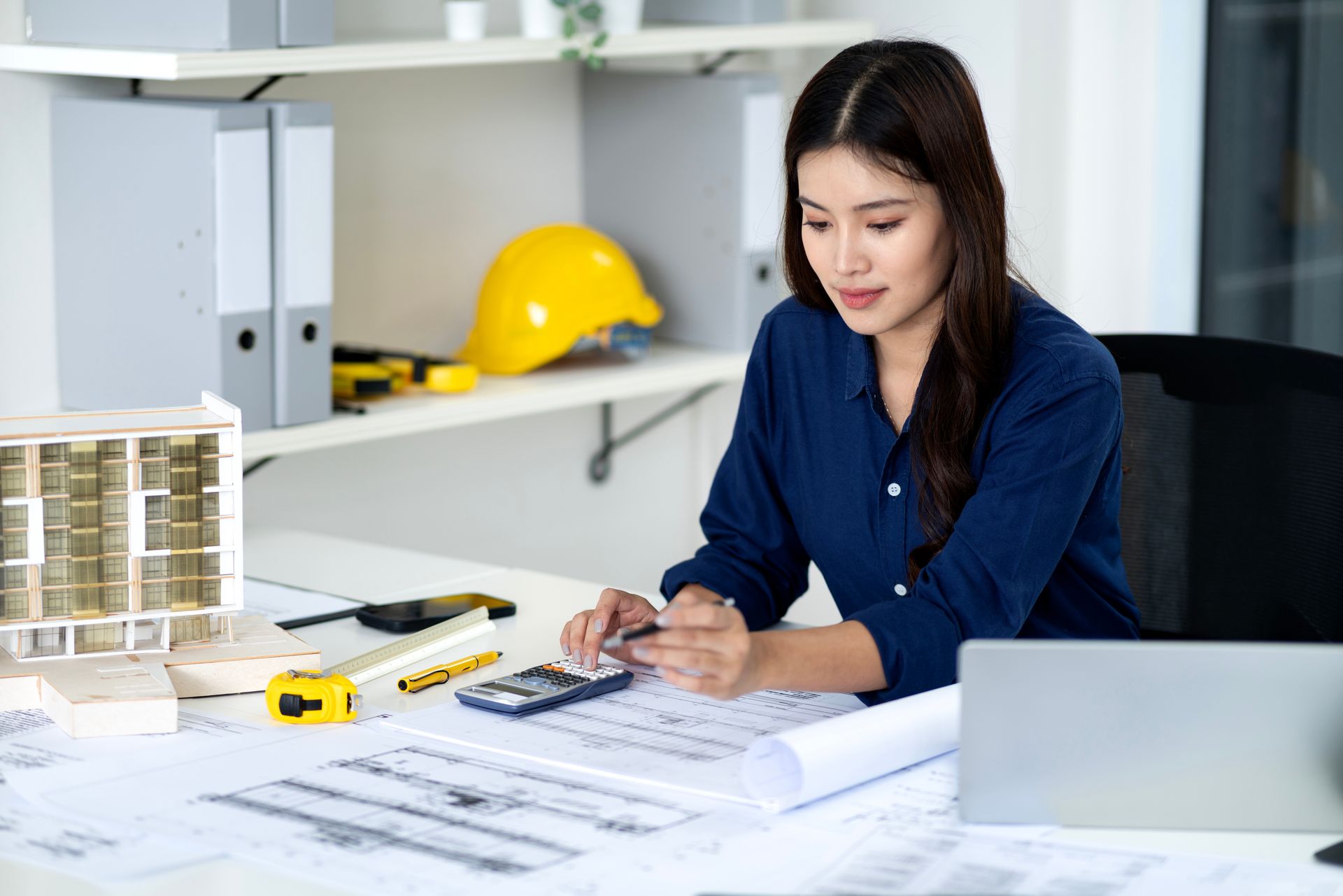 Architect reviewing blueprints at a desk with a yellow hard hat, laptop, and building model nearby