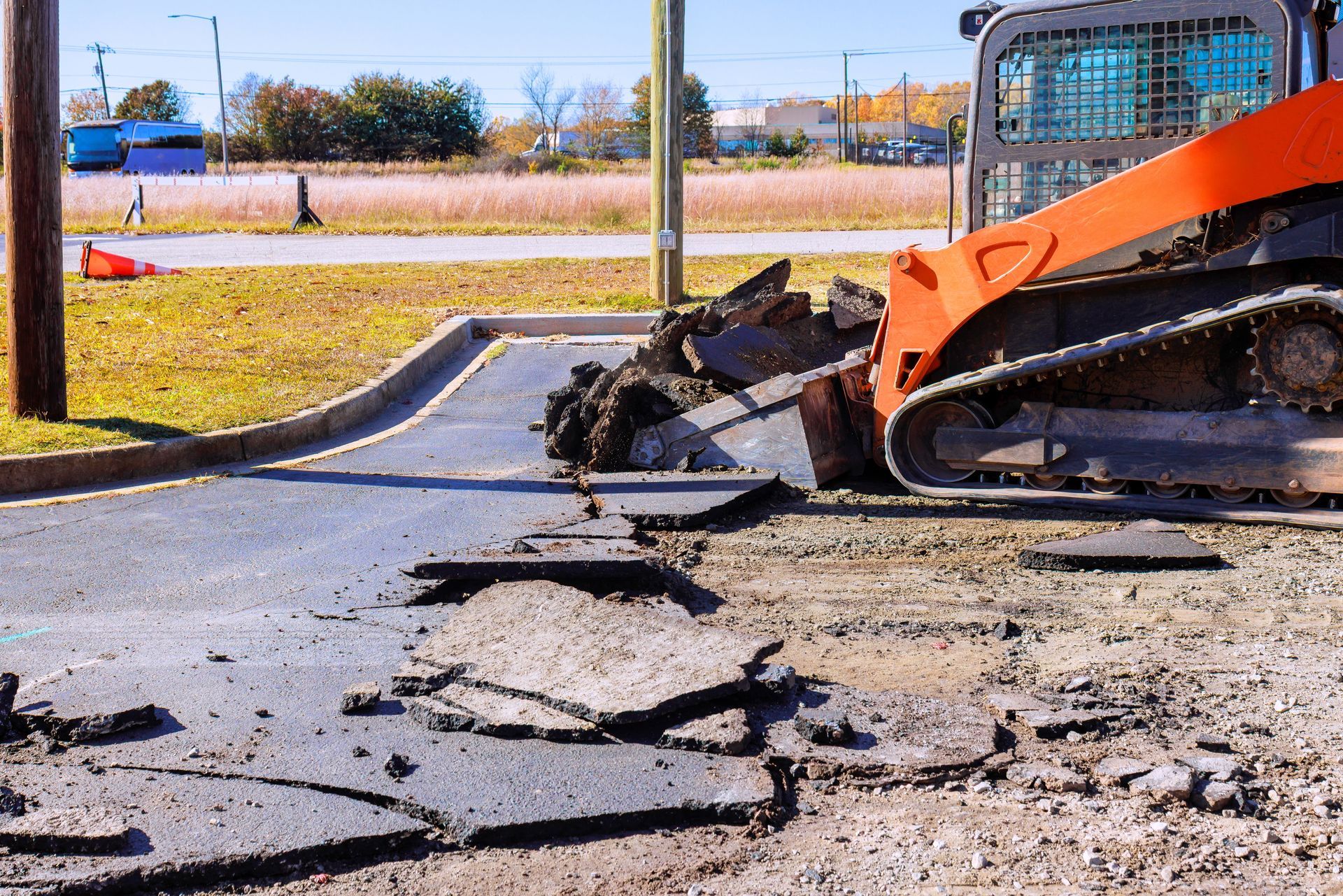 Orange skid-steer breaking up cracked pavement on a roadside construction site