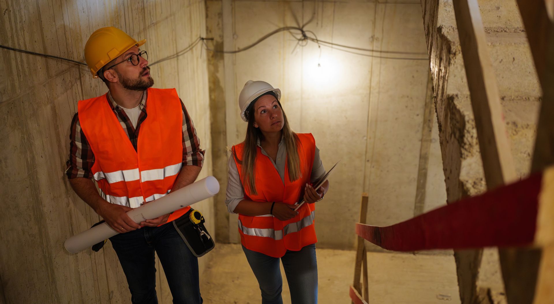 Two construction workers in hard hats and orange vests examine plans inside a tunnel or shaft.