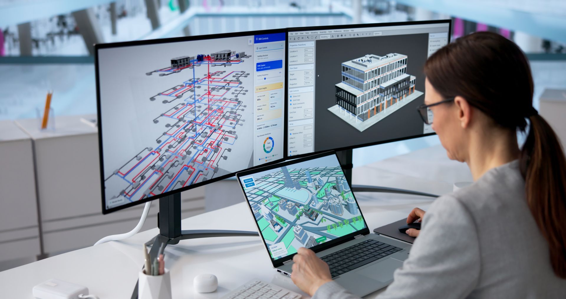 Person reviewing technical drawings on dual monitors at a desk, with a laptop showing a 3D building model.
