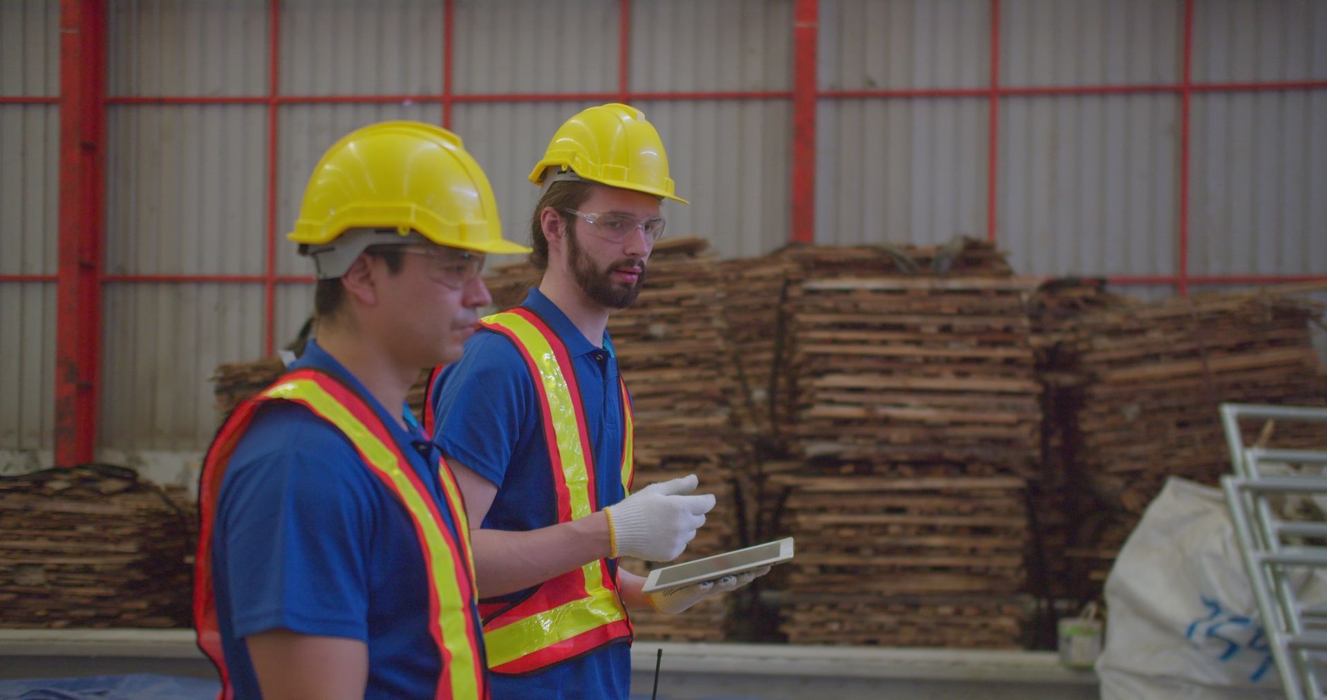 Two workers in hard hats and safety vests stand in a warehouse with stacked lumber.