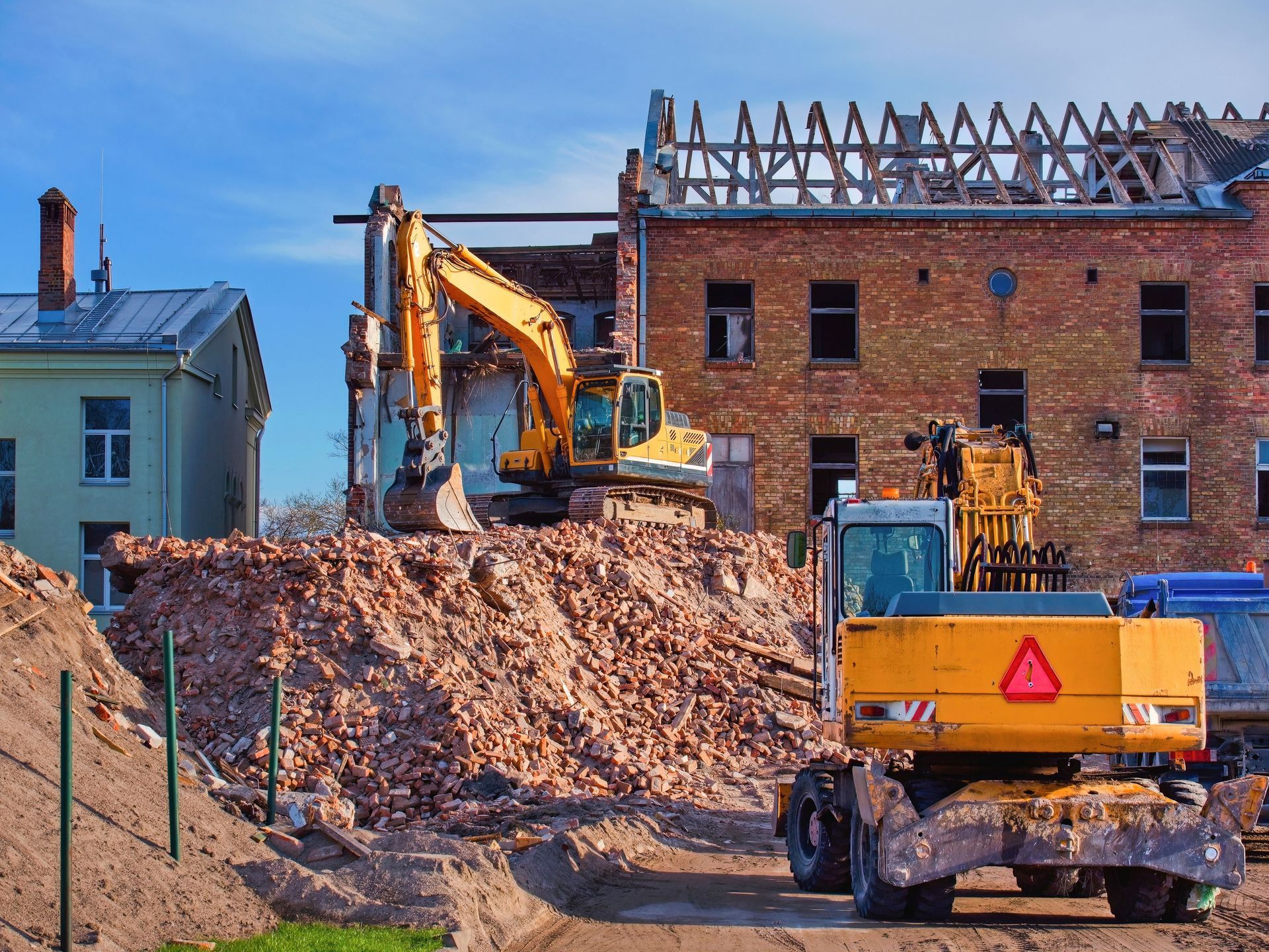 Construction site with excavator and yellow dump truck beside a brick building and dirt pile