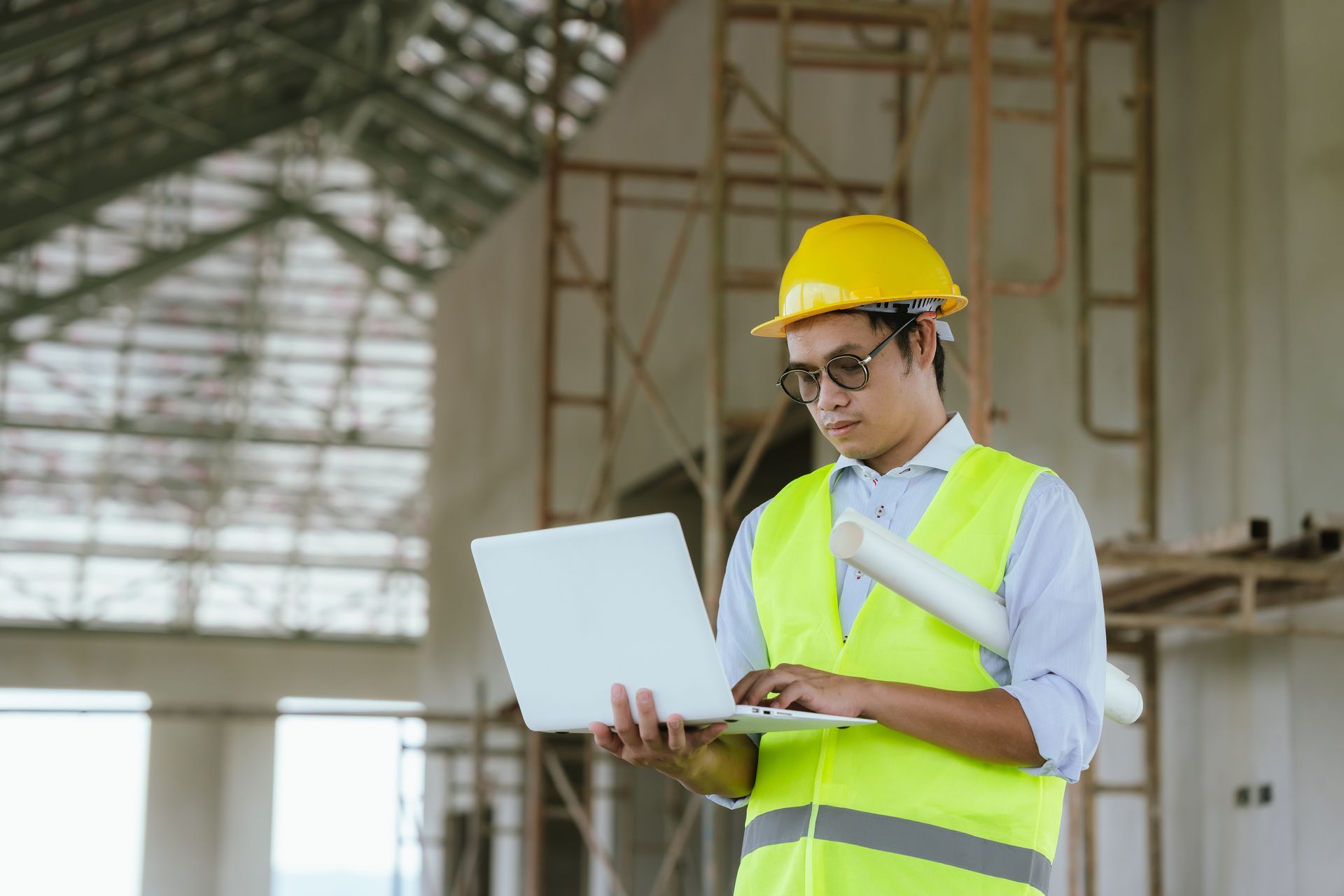 Worker in yellow safety vest and hard hat using a laptop at a construction site.